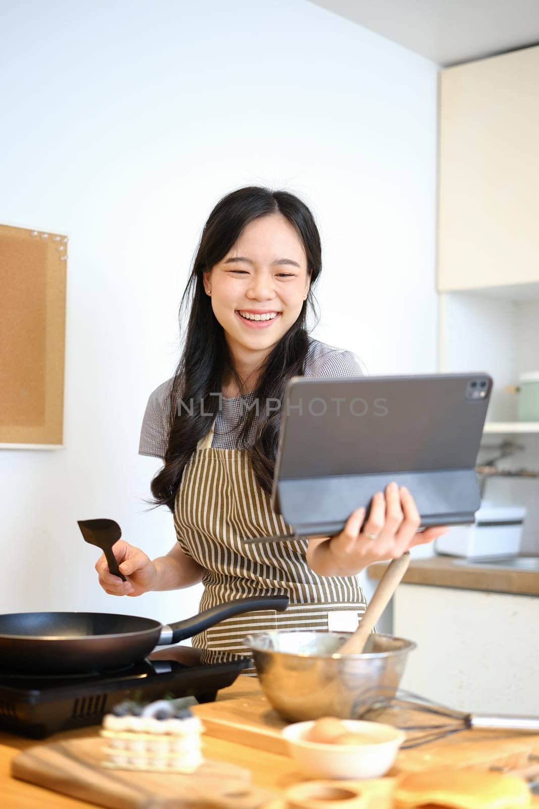 Cheerful asian housewife cooking at home and following an online recipe on digital tablet. People, technology and lifestyle.