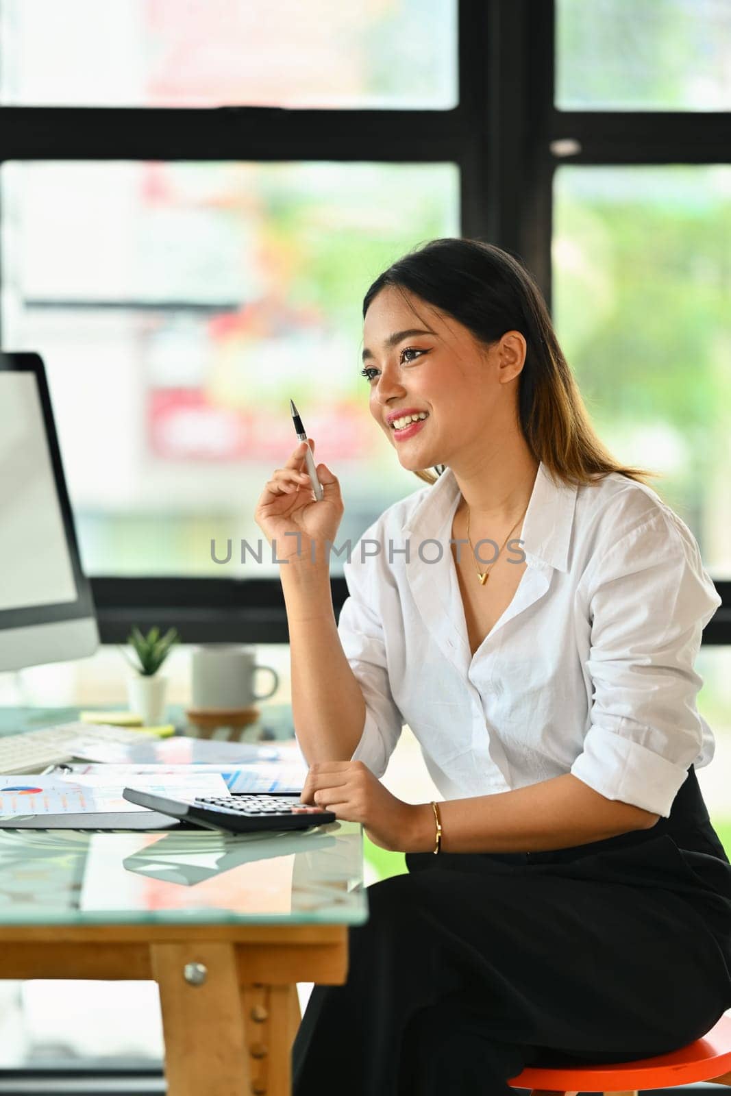 Beautiful female accountant sitting at desk and looking away thoughtfully, pondering about business strategy.