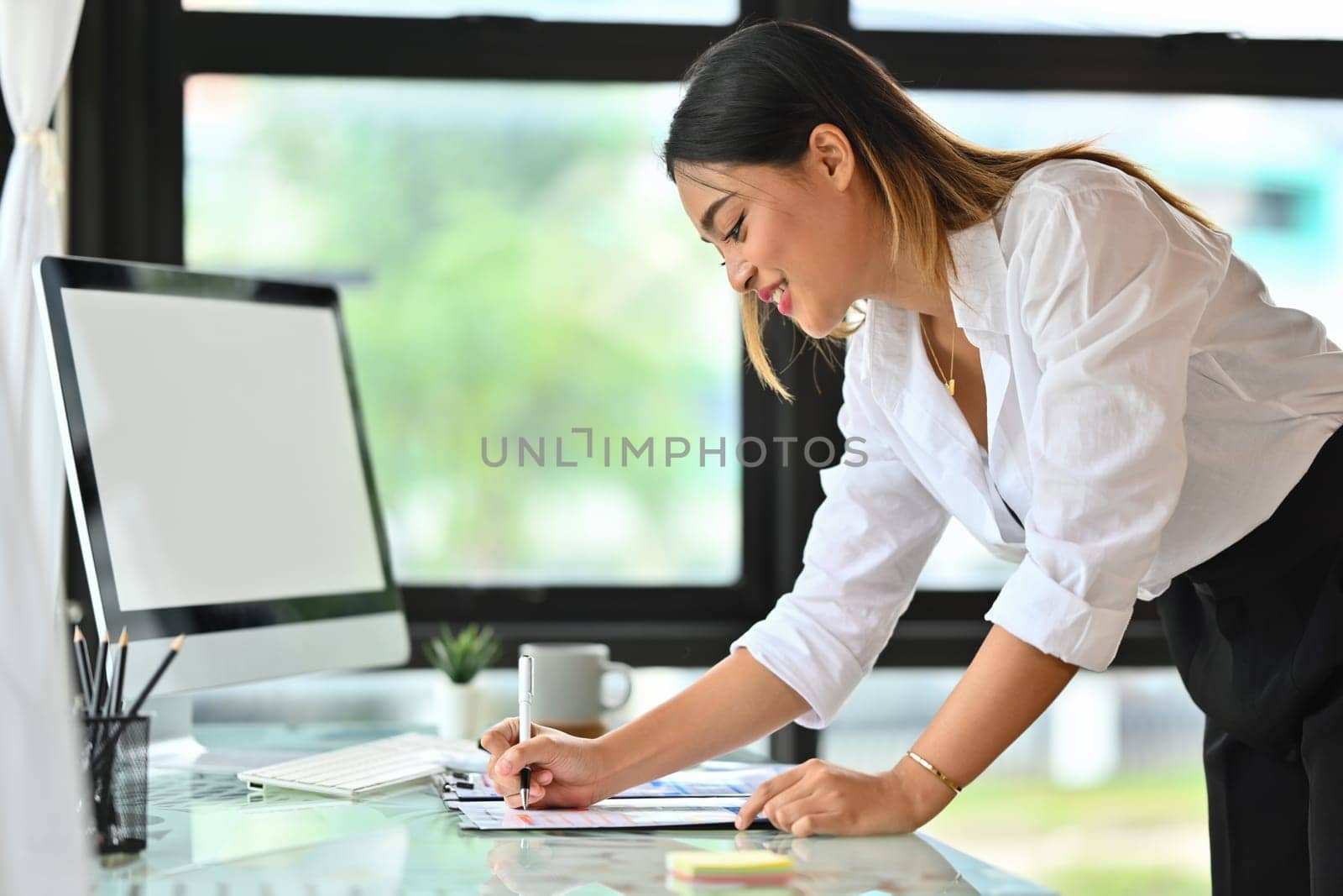 Charming millennial female manager standing at her workplace and writing on paperwork.