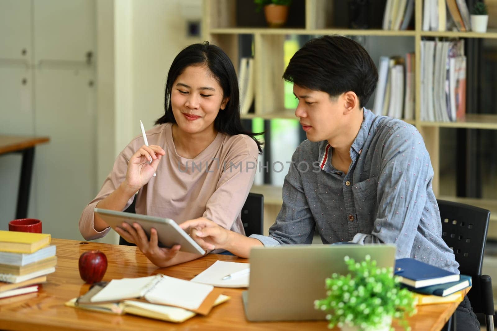 Two college students using digital tablet in library, studying together for exam or research for project.