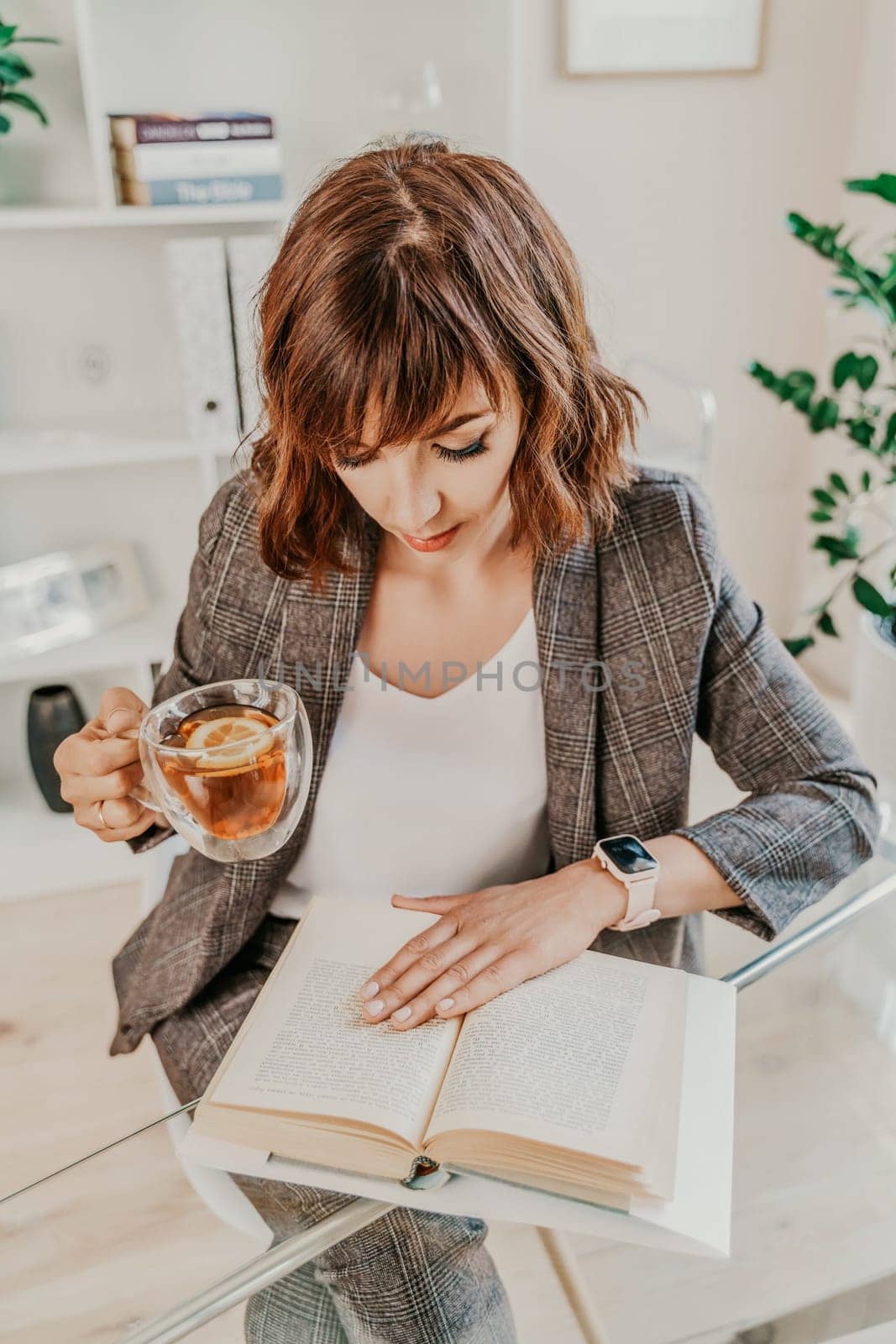 Woman tea office. A young business woman is drinking tea and reading a book while sitting at a table in the office. Dressed in a business suit and white blouse. by Matiunina