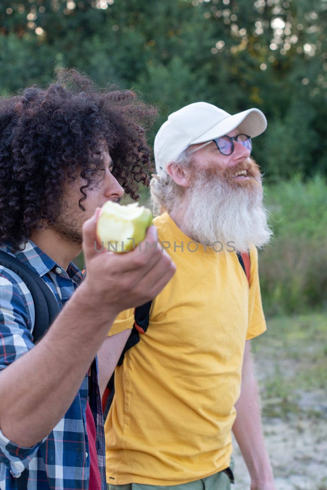 two friends of different ages go hiking with backpacks, and an Arab guy with black hair eats a green apple with appetiteagainst the background of a forest, High quality photo