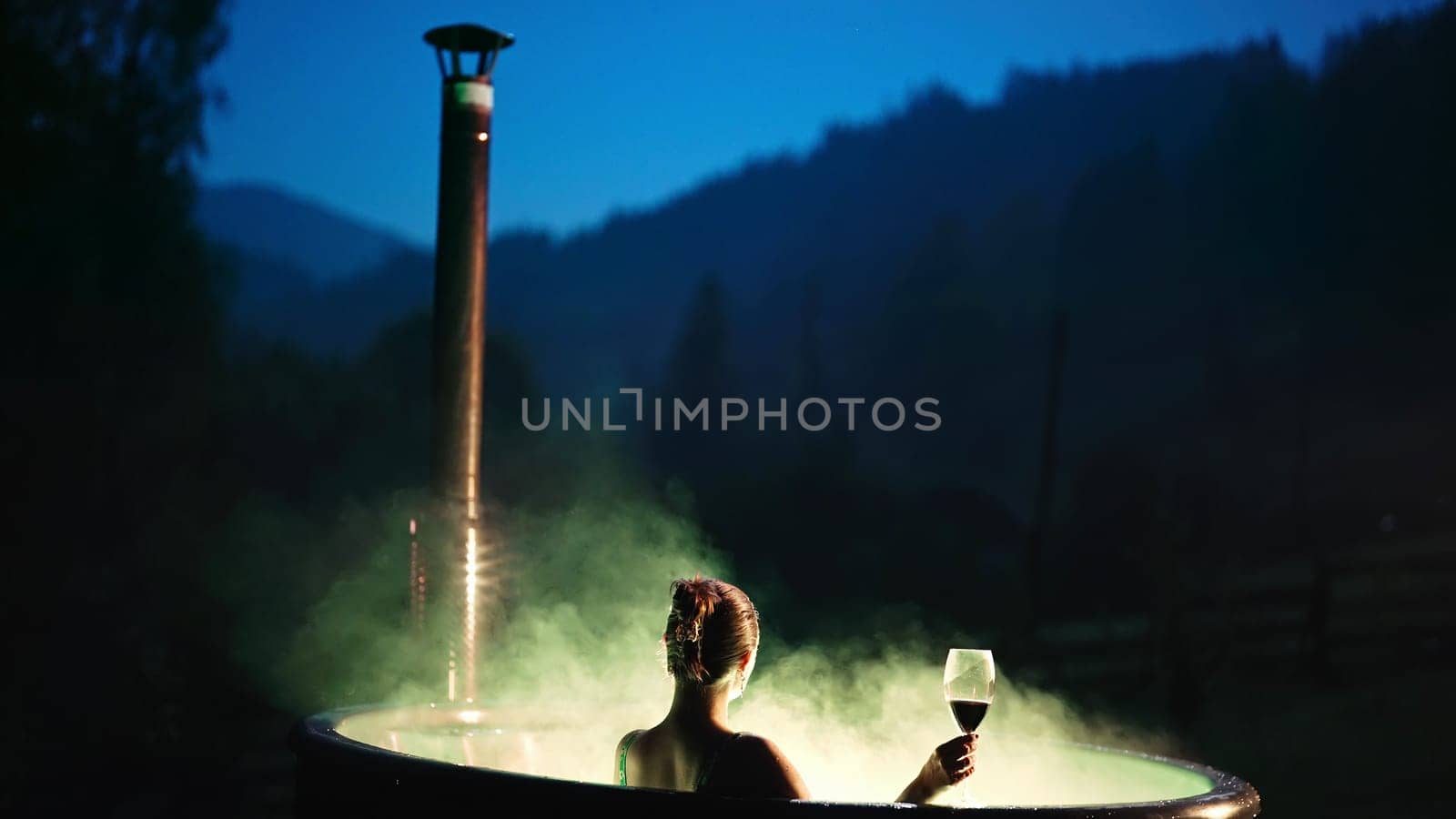 Woman spending holidays resting on wild nature, drinks red wine. Lady in hot wooden vat heated by firewood, bathing with mineral water. Aestethic. Eco living, wellness center, hydrotherapy.
