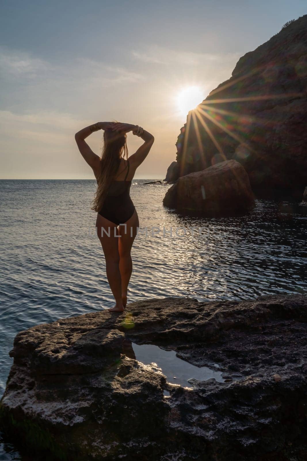 Woman tourist enjoying the sunset over the sea mountain landscape. Sits outdoors on a rock above the sea. She is wearing jeans and a blue hoodie. Healthy lifestyle, harmony and meditation.