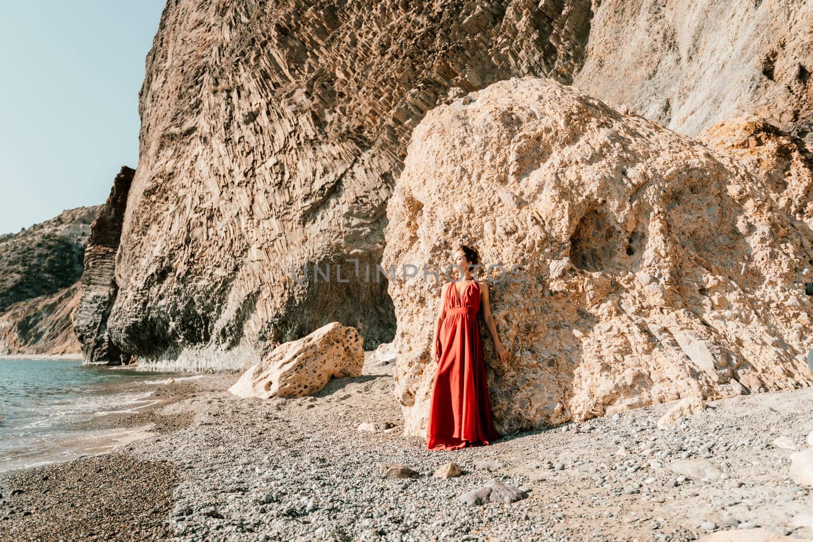 Woman red dress sea. Woman in a long red dress posing on a beach with rocks on sunny day. Girl on the nature on blue sky background
