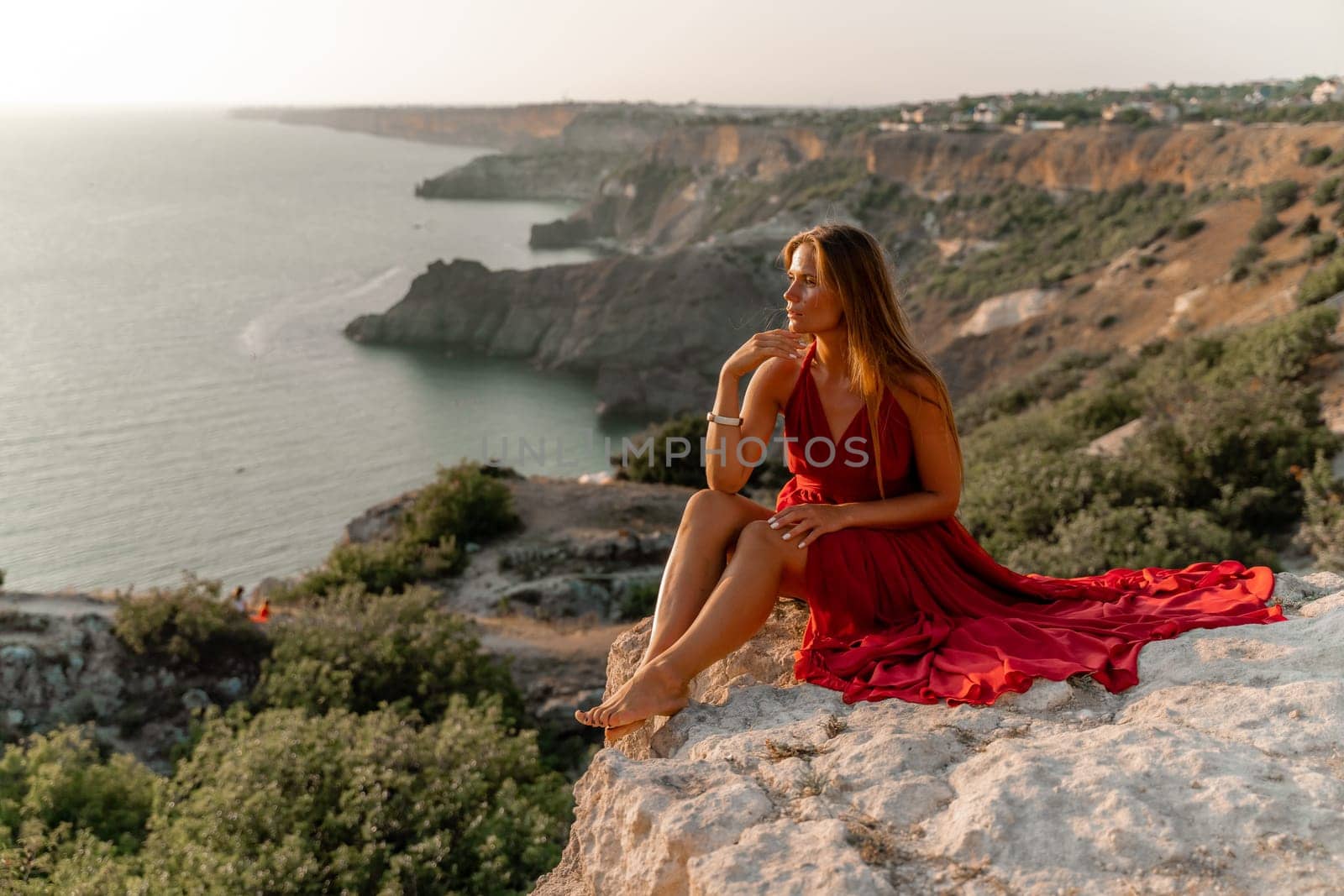 Woman sunset sea red dress, side view a happy beautiful sensual woman in a red long dress posing on a rock high above the sea on sunset