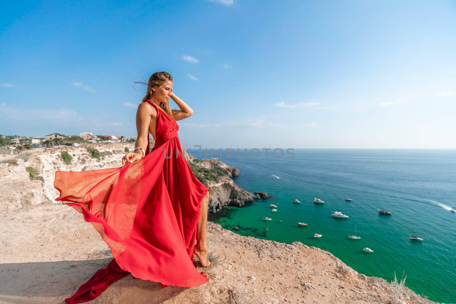 Woman sea red dress yachts. A beautiful woman in a red dress poses on a cliff overlooking the sea on a sunny day. Boats and yachts dot the background