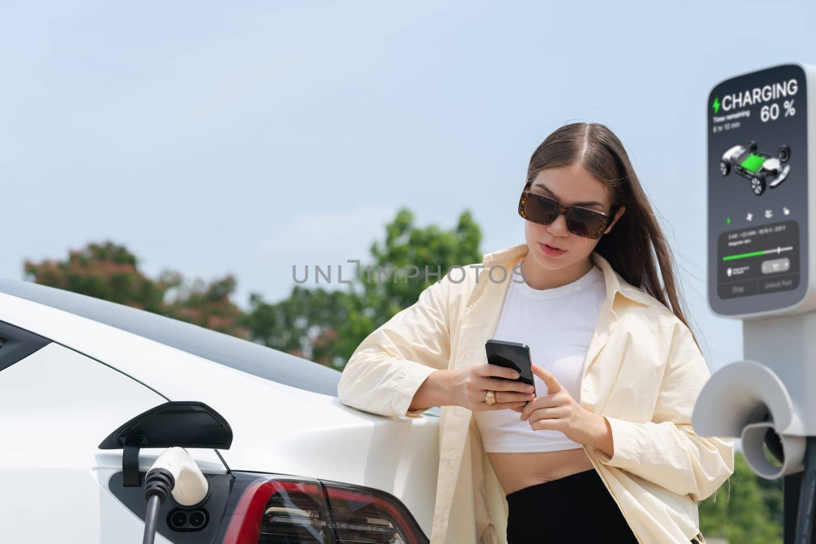 Young woman use smartphone to pay for electricity at public EV car charging station green city park. Modern environmental and sustainable urban lifestyle with EV vehicle. Expedient