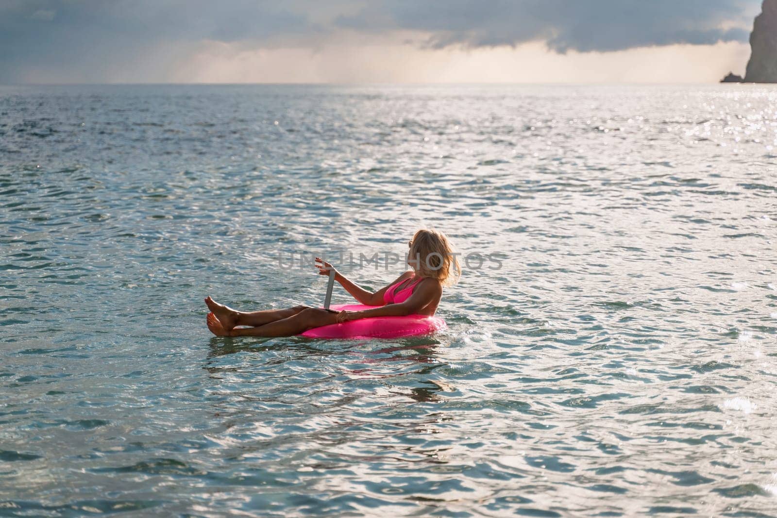 Woman works on laptop in sea. Freelancer, blond woman in sunglases floating on an inflatable big pink donut with a laptop in the sea at sunset. People summer vacation rest lifestyle concept