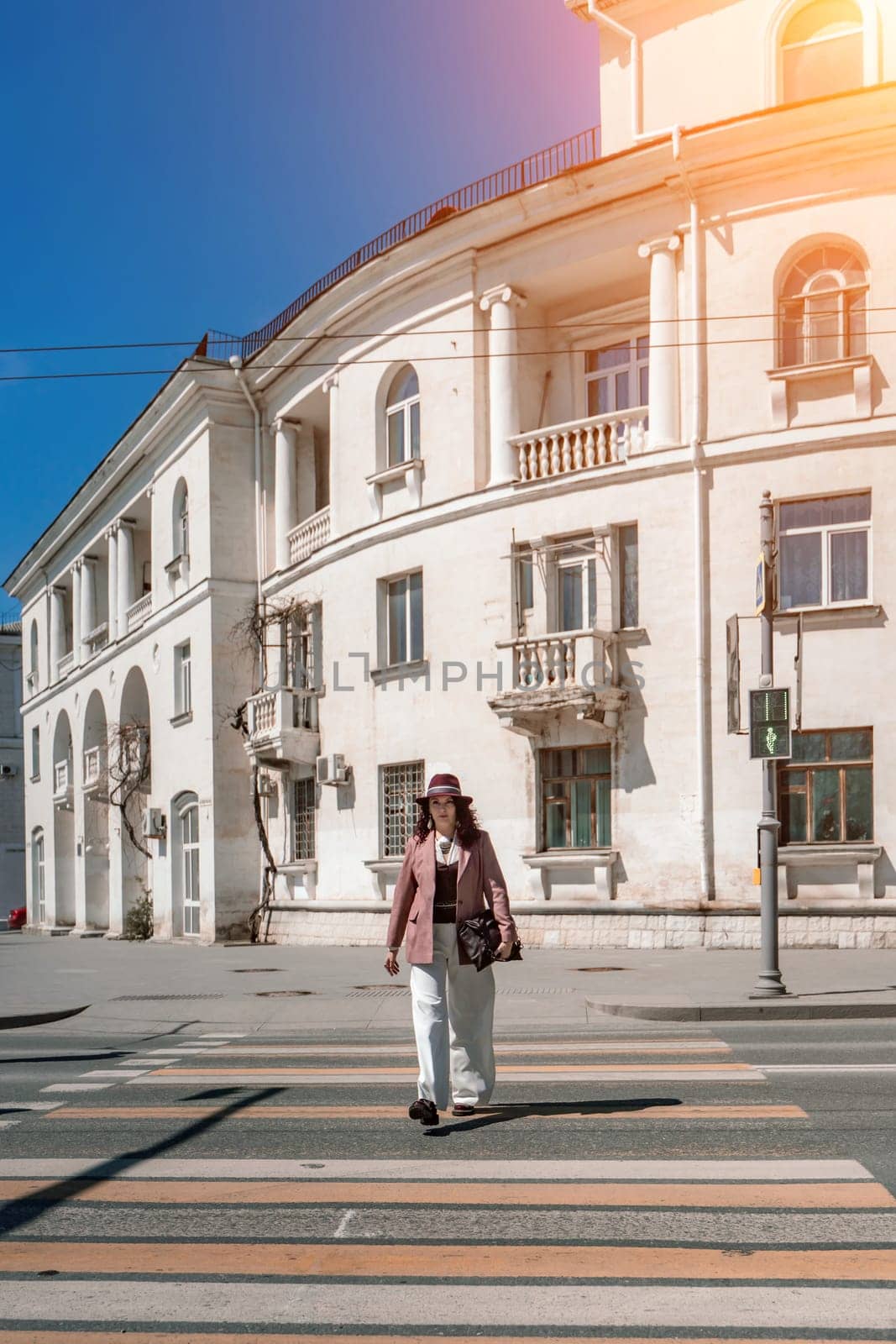 Woman city road crossing. Stylish woman in a hat crosses the road at a pedestrian crossing in the city. Dressed in white trousers and a jacket with a bag in her hands. by Matiunina