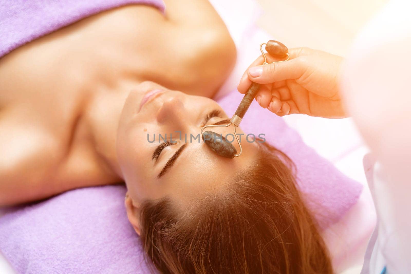 Facial massage. Side view european woman getting massage with jade face roller gouache in spa salon.