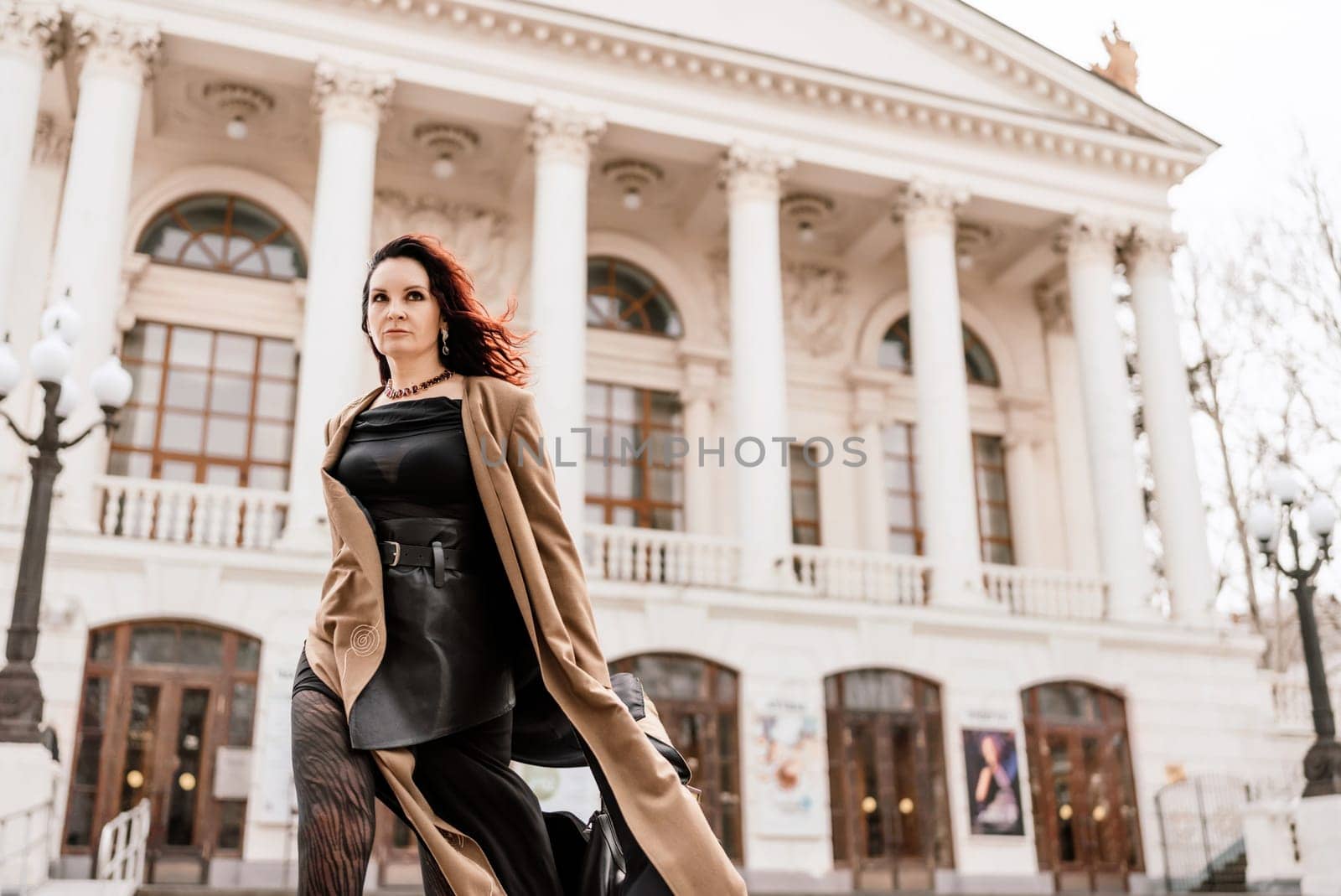 Woman street lifestyle. Image of stylish woman walking through European city on sunny day. Pretty woman with dark flowing hair, dressed in a beige raincoat and black, walks along the building. by Matiunina