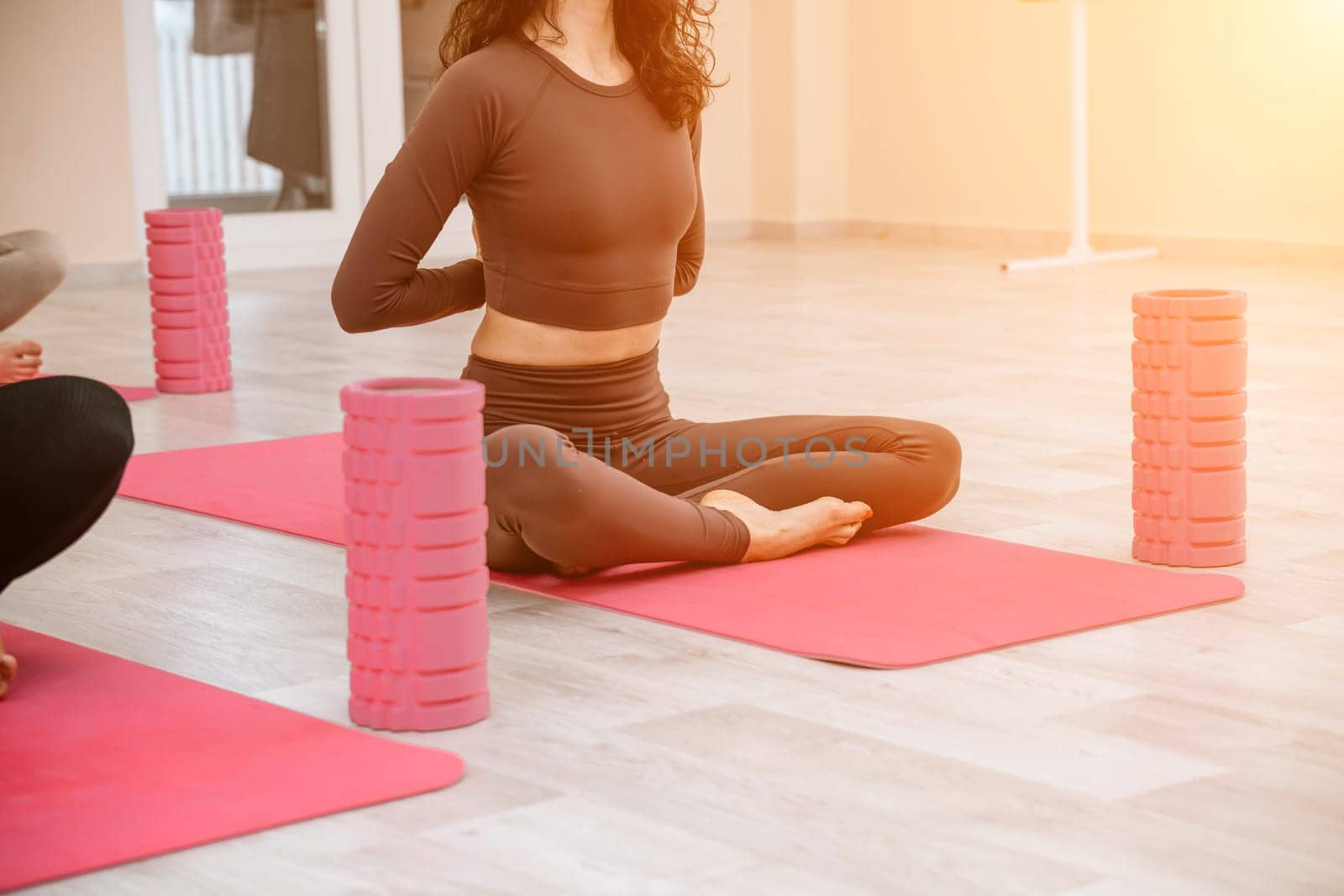 A group of six athletic women doing pilates or yoga on pink mats in front of a window in a beige loft studio interior. Teamwork, good mood and healthy lifestyle concept