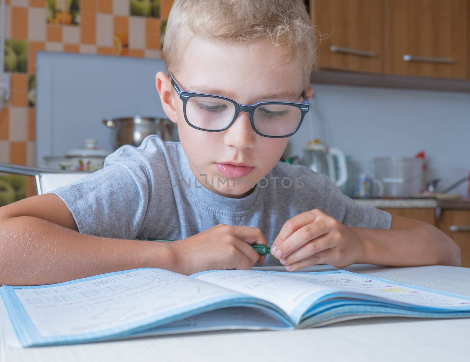 Tired sad little child boy sits at a table in the kitchen and writes homework. Boring lessons.