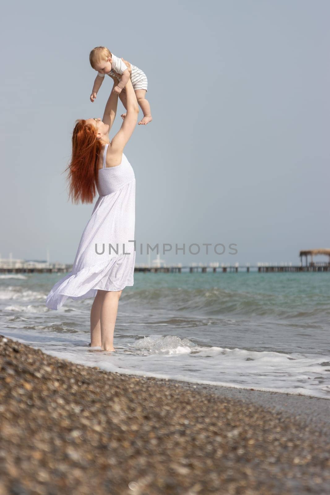 Happy woman raising up her little son standing on the beach above the sea. Mid shot