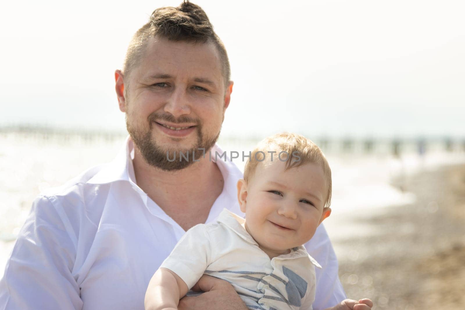 Happy father on vacation - smiling man standing on a beach holding his little baby son. Portrait