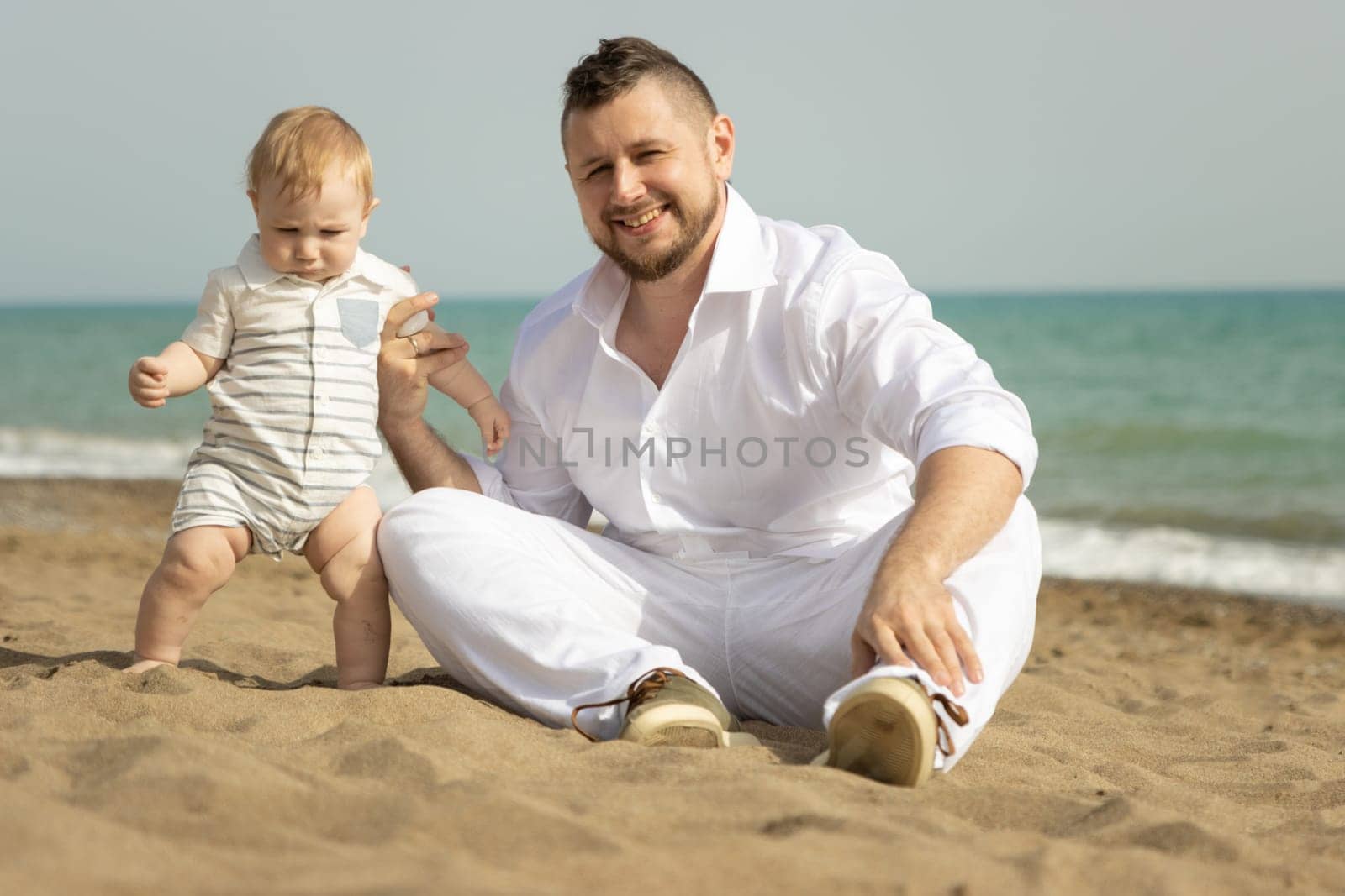Happy father sitting on a beach with his little son. Mid shot