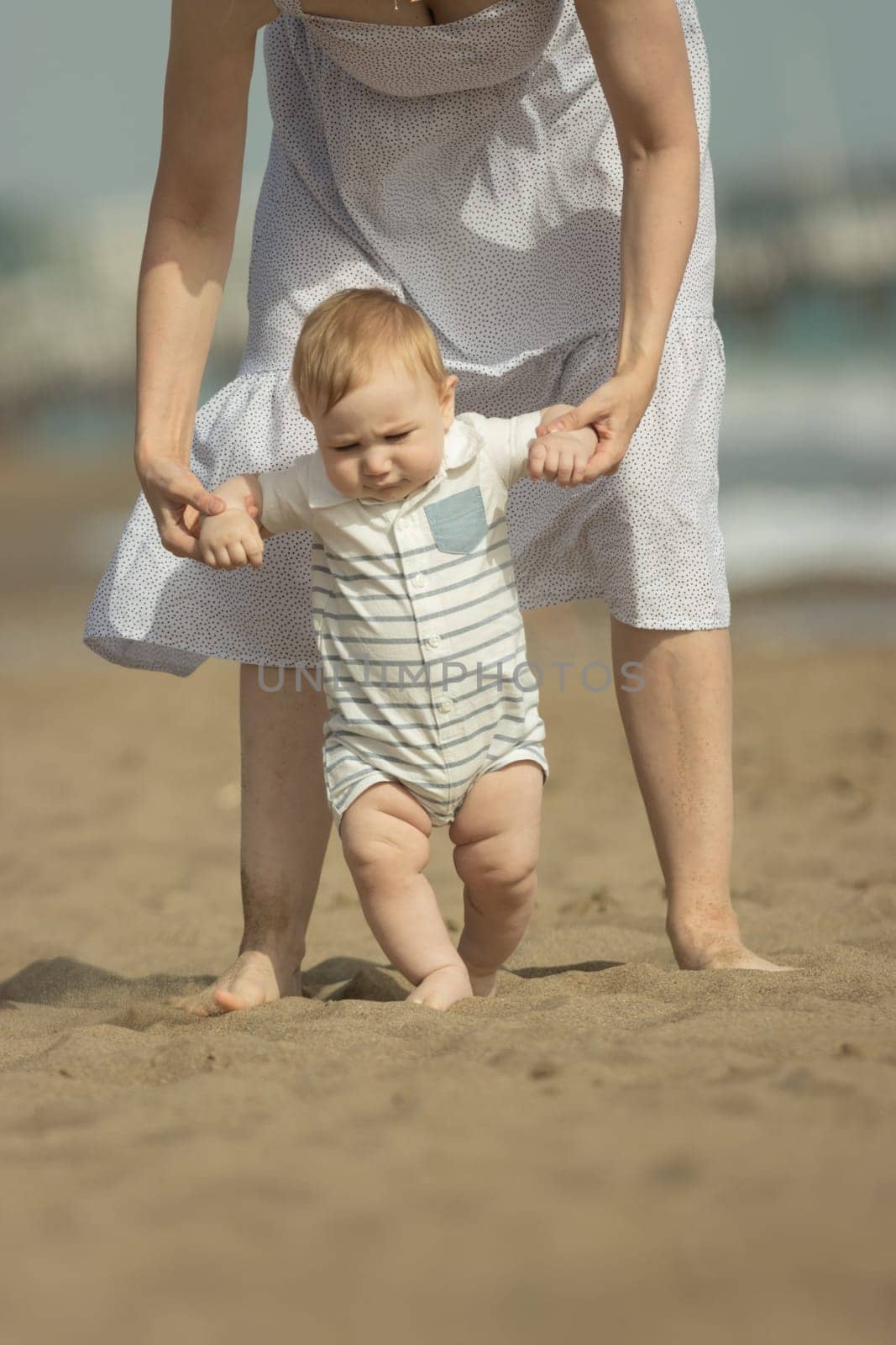 A little boy learns how to walk on the sand with support of his mother. Mid shot