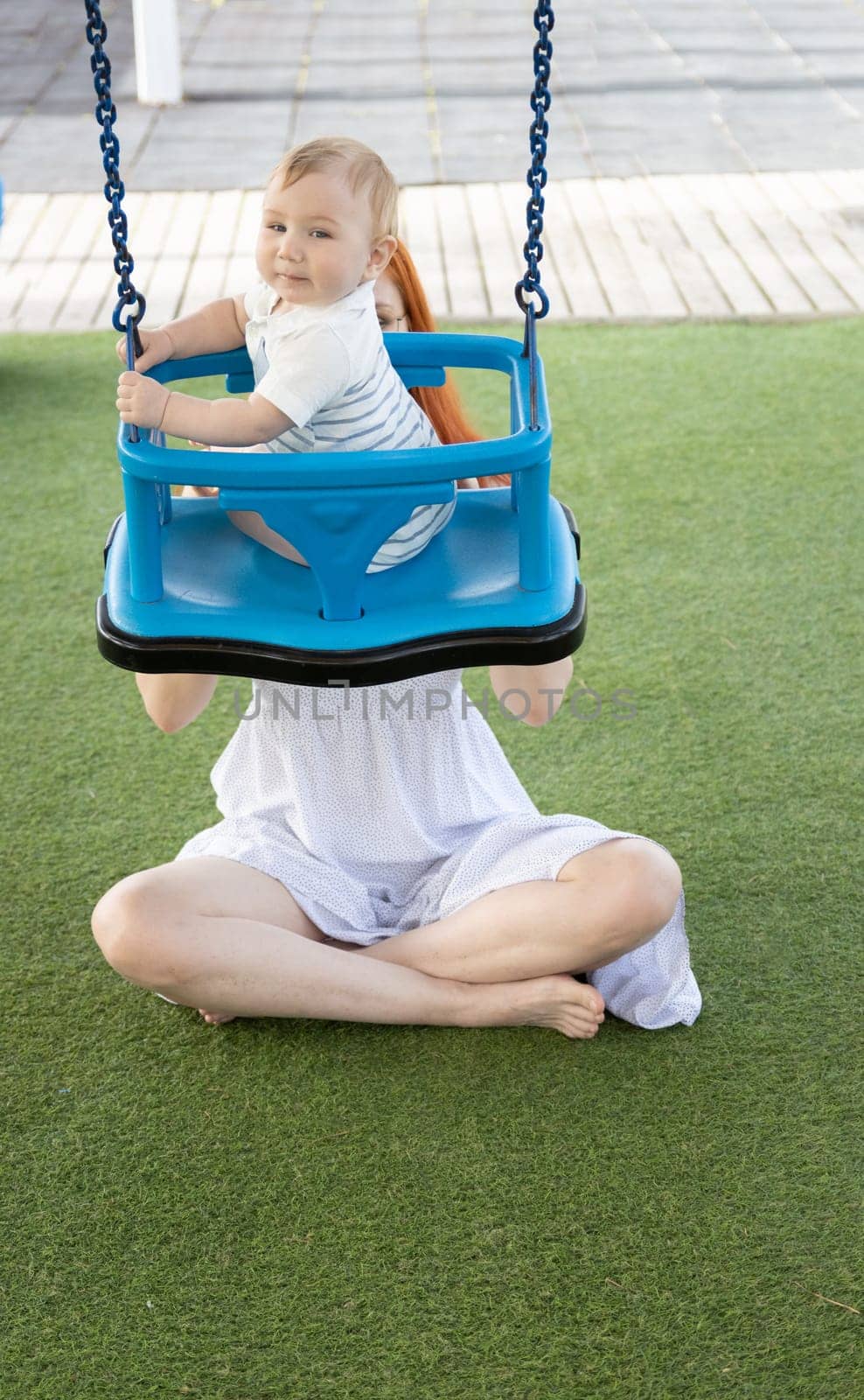 Young mom shakes her young son on a swing in the playground by Studia72