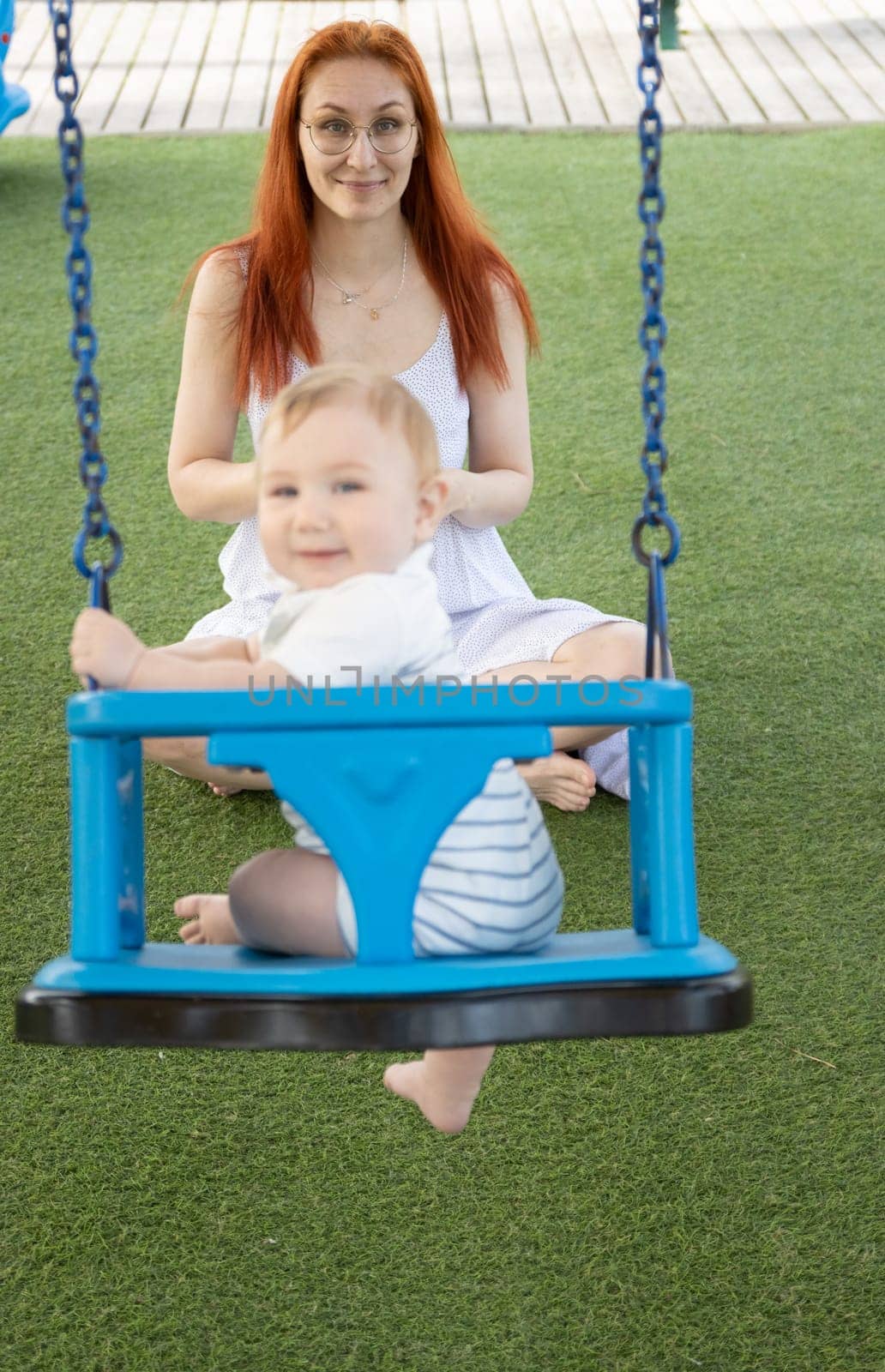 A little funny boy on swings on an outside playground with his ginger mother - looking in the camera. Mid shot