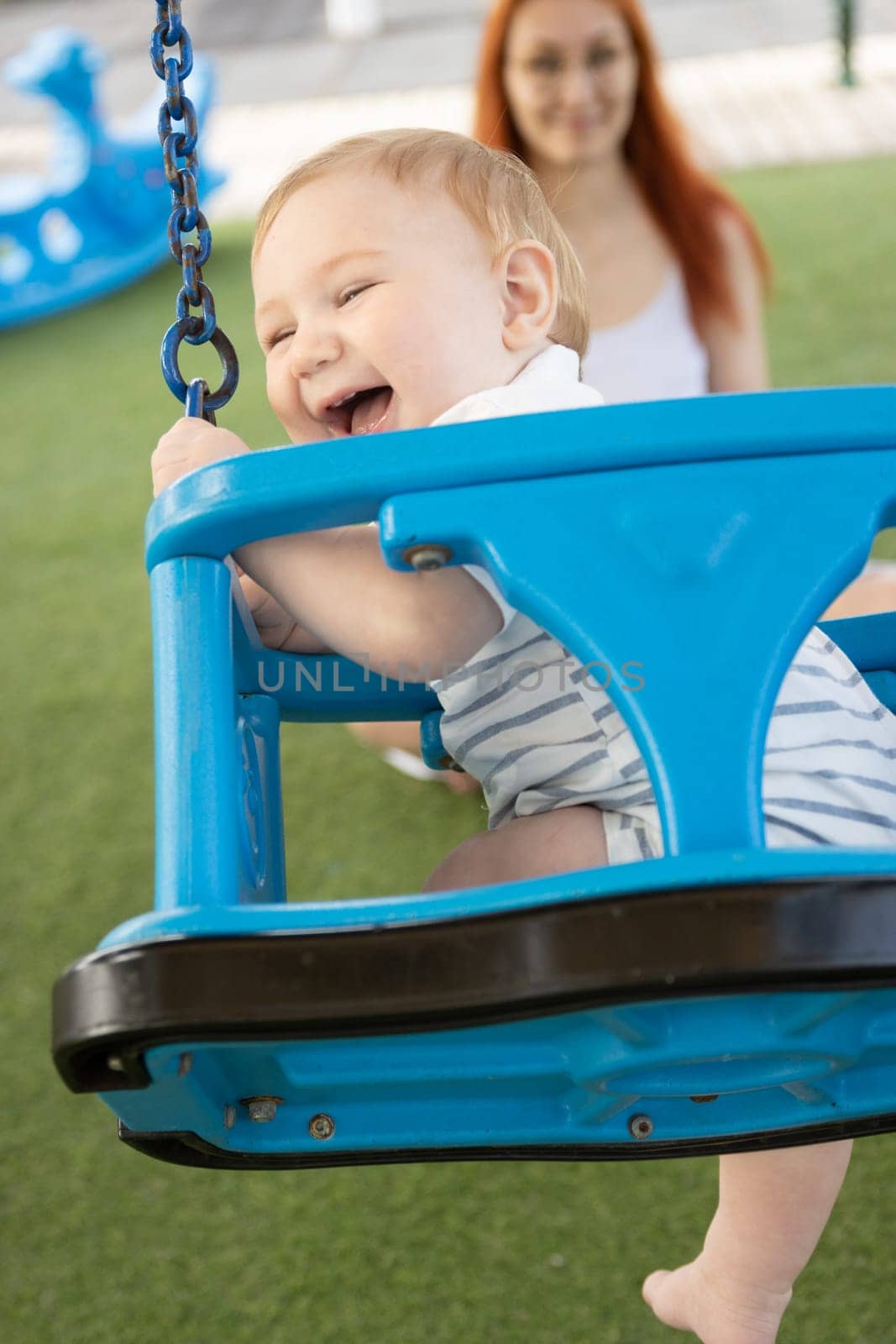 A little laughing boy on swings on an outside playground with his ginger mother - looking in the camera. Mid shot