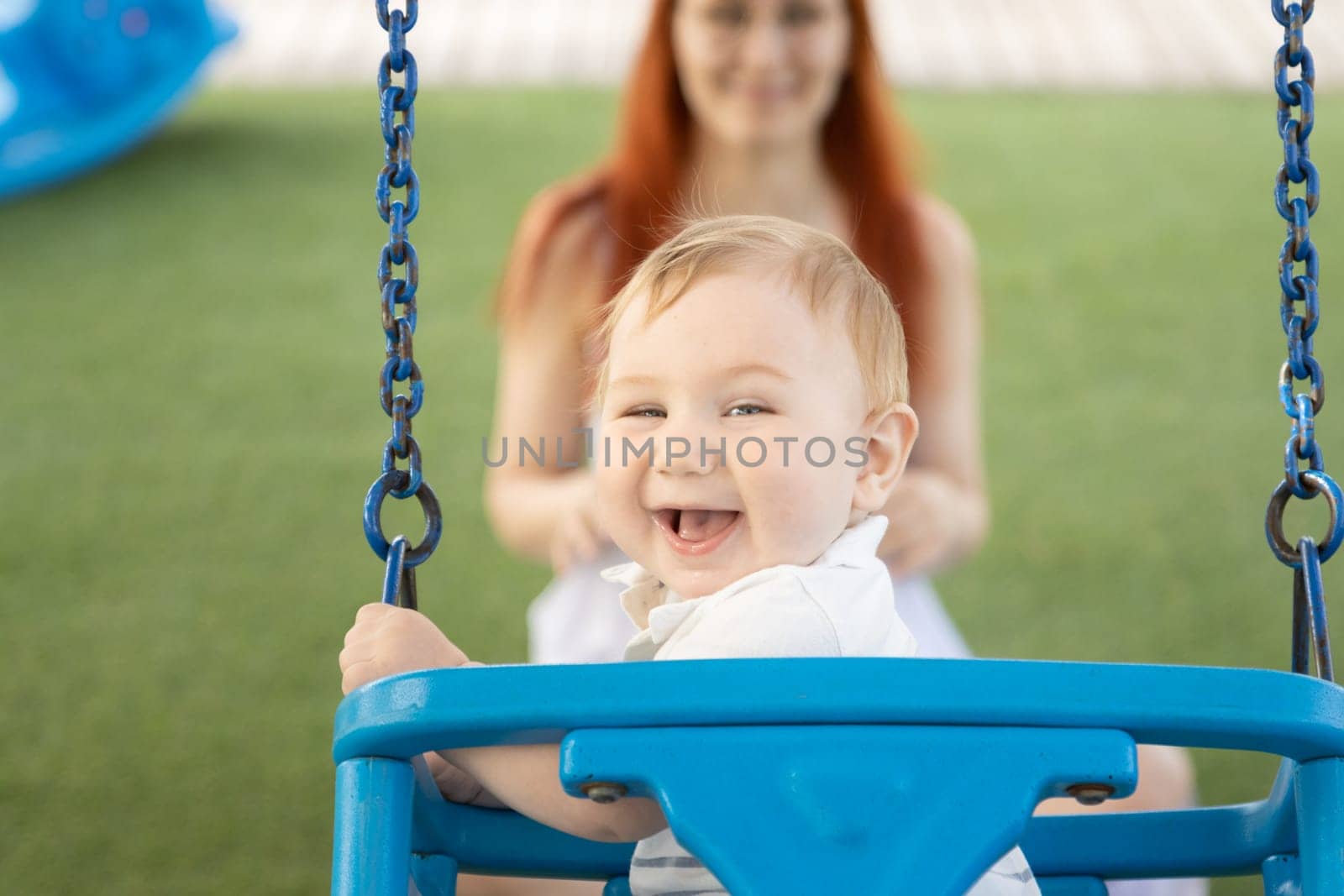 A little laughing baby on swings on an outside playground with his ginger mother by Studia72