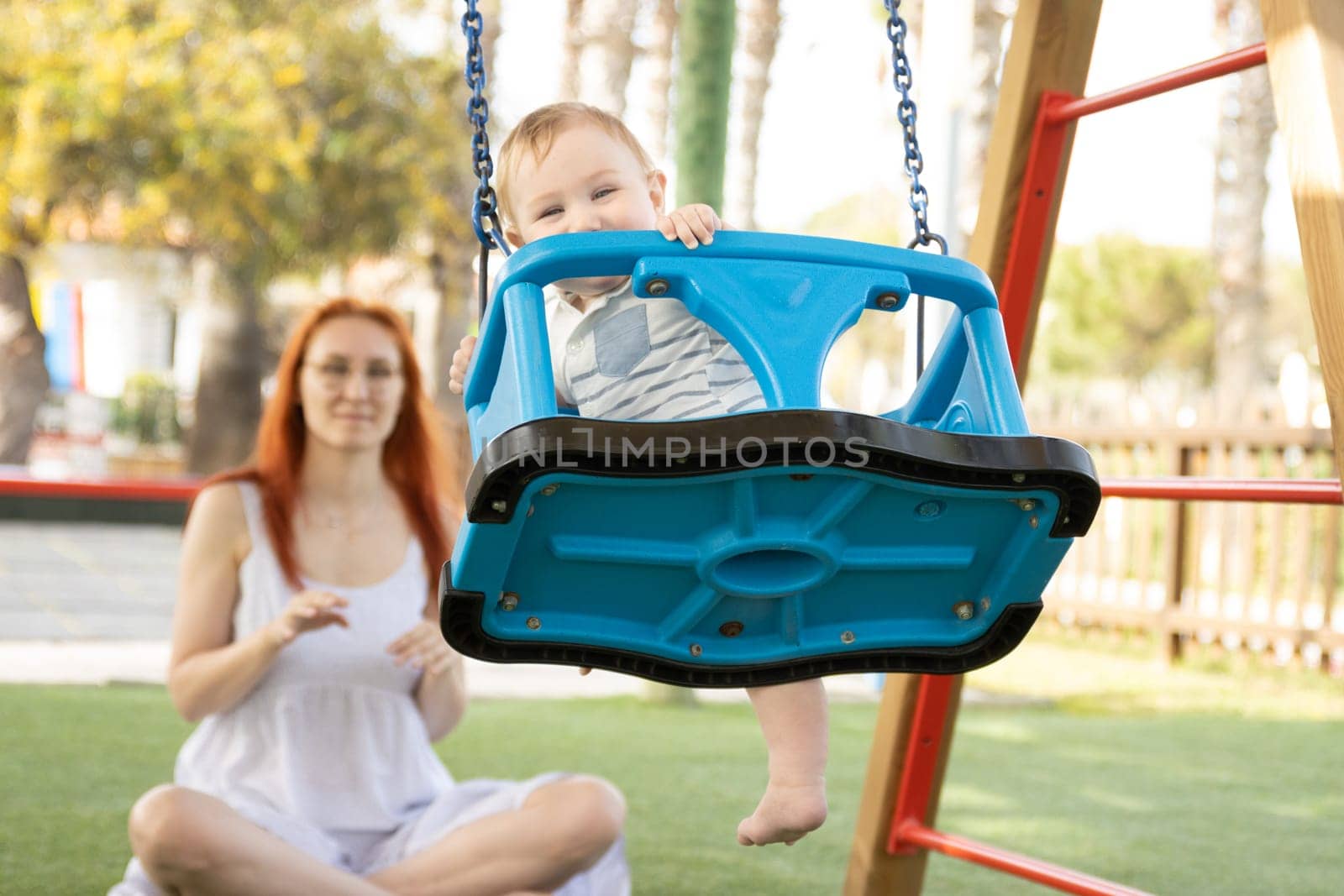 A little funny baby on swings on an outside playground with his ginger mother. Mid shot
