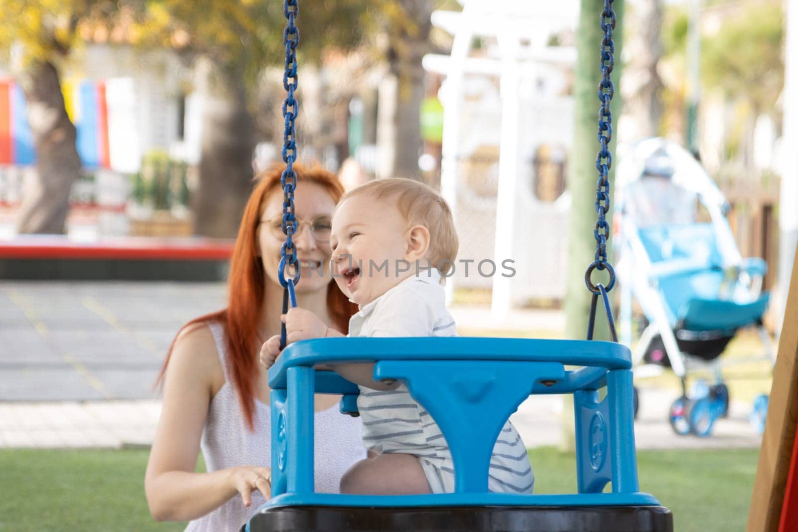 Happy family vacation - funny baby swings on swings in playground and his mom sits near him. Mid shot