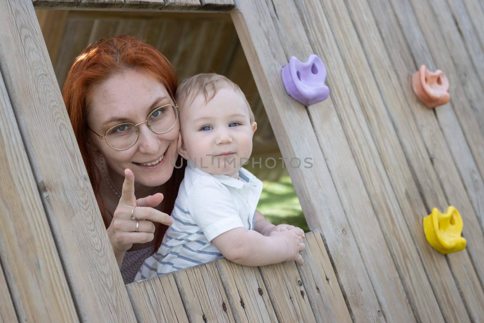 Little baby and his mother sits in a wooden house on the playground - looking in the camera. Mid shot