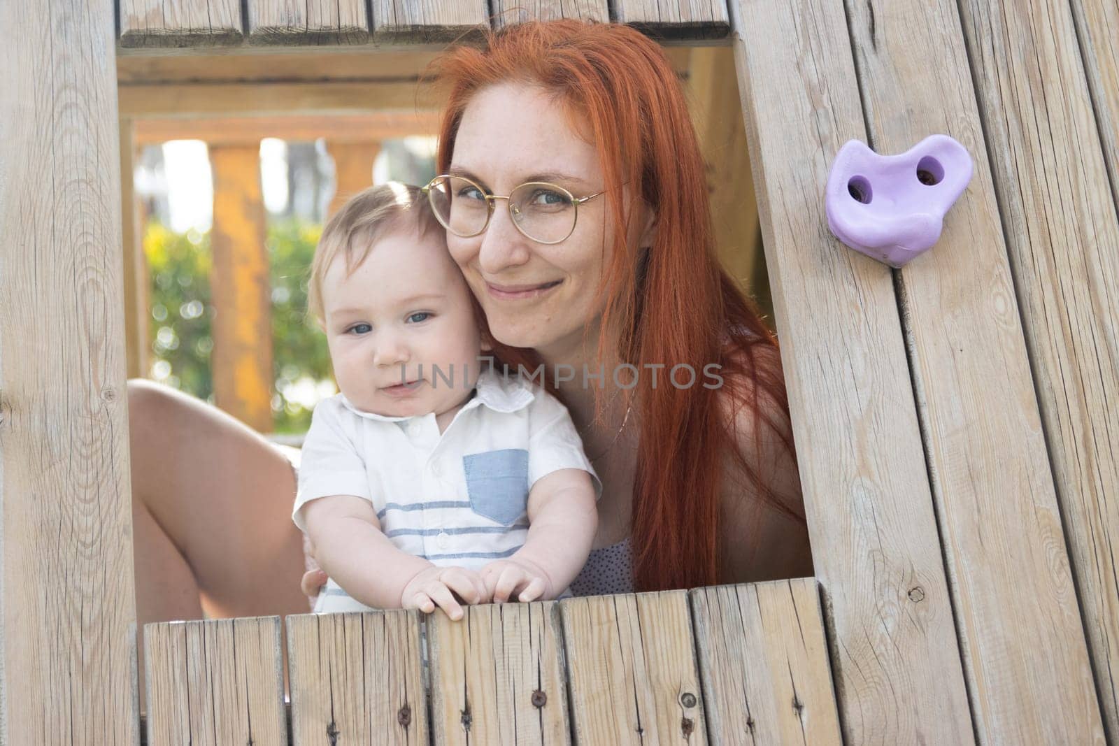 Cute baby and his mother sits in a wooden house on the playground - looking in the camera by Studia72
