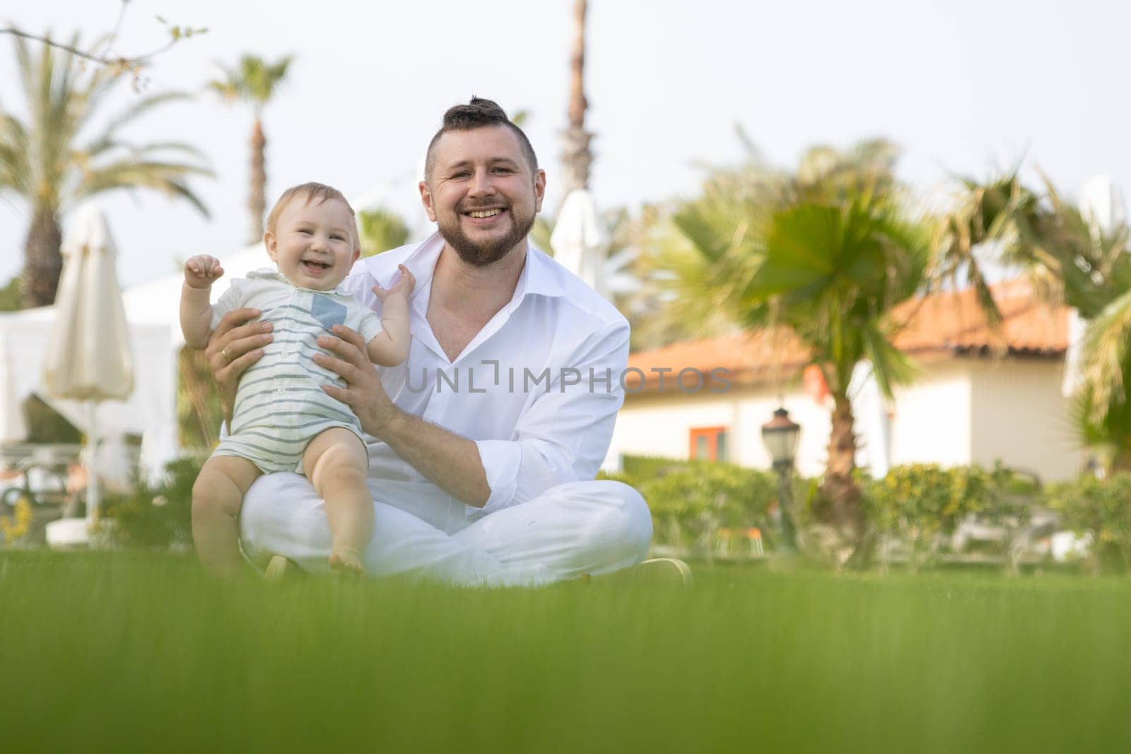 Happy father and his son sit on the grass - looking into the camera. Mid shot