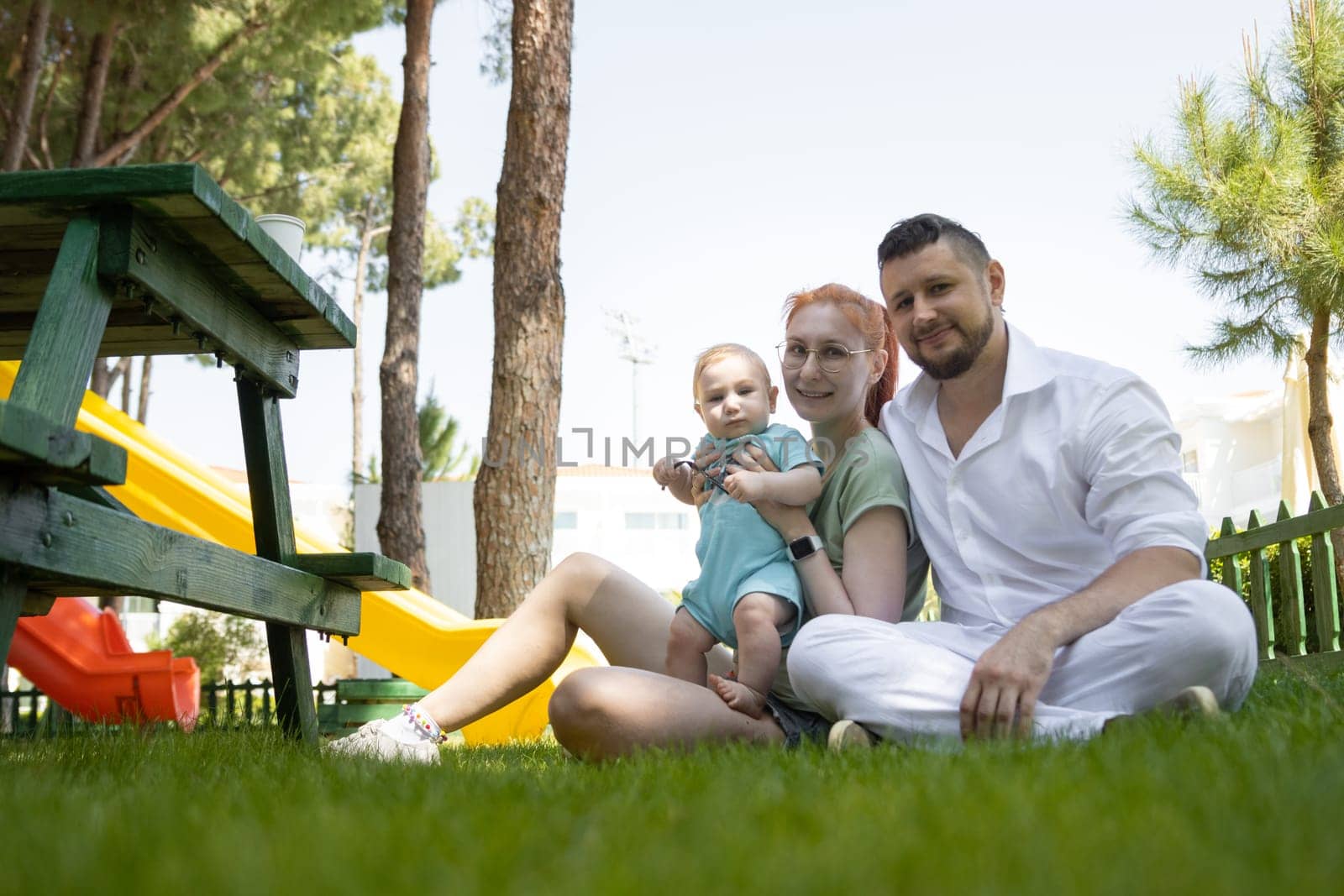 Happy parents and their cute son sit on the grass - looking into the camera. Mid shot