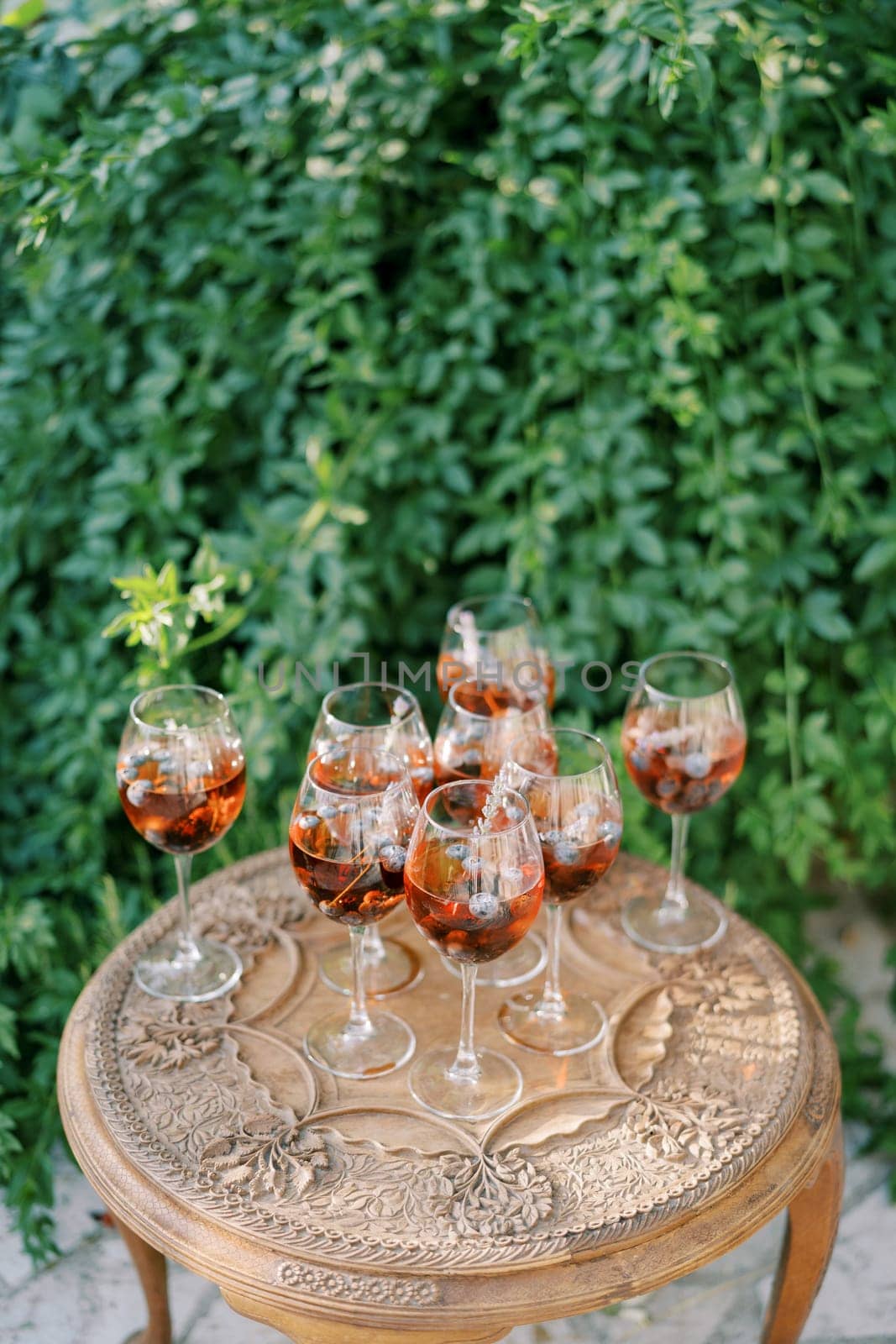 Glasses with red wine and floating grapes inside stand on a carved wooden table in the garden by Nadtochiy