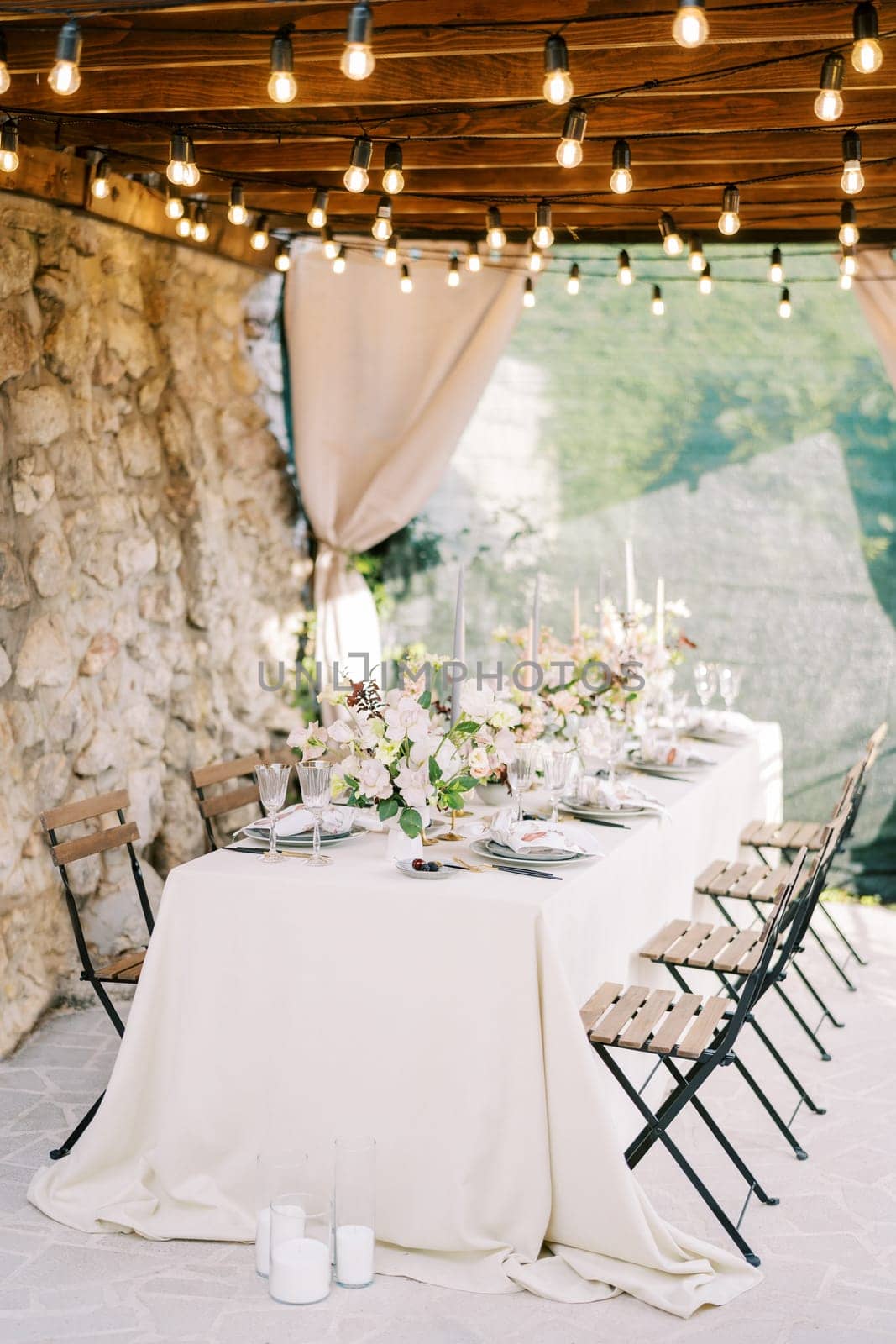 Laid festive table with chairs stands against a stone wall under a wooden canopy with glowing garlands. High quality photo
