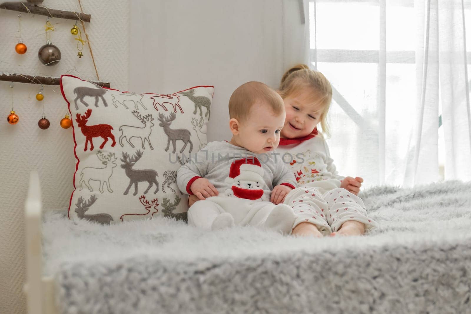 Brother and sister in Christmas pajamas are sitting on the bed with decorations.