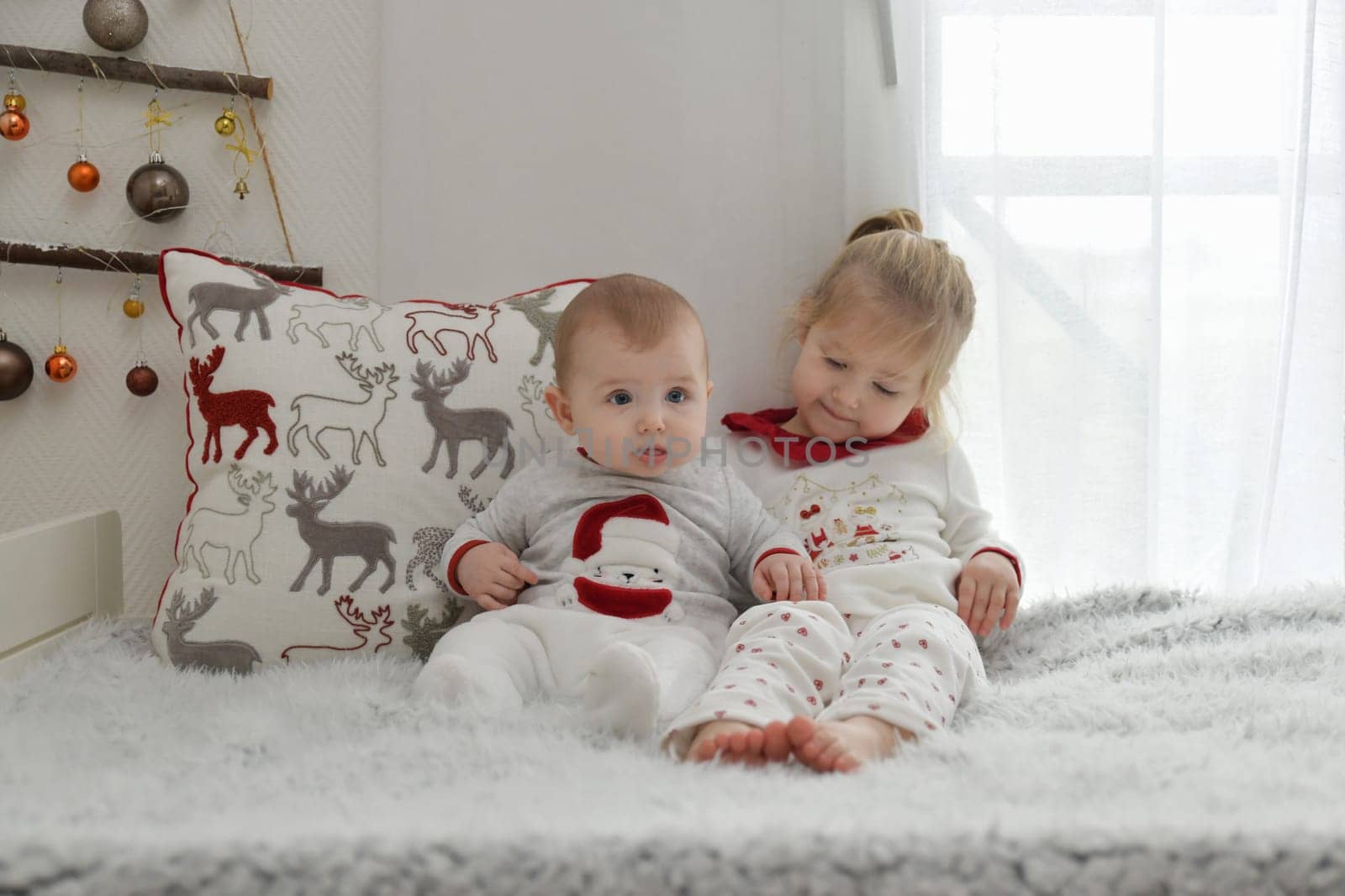 Brother and sister in Christmas pajamas are sitting on the bed with decorations.