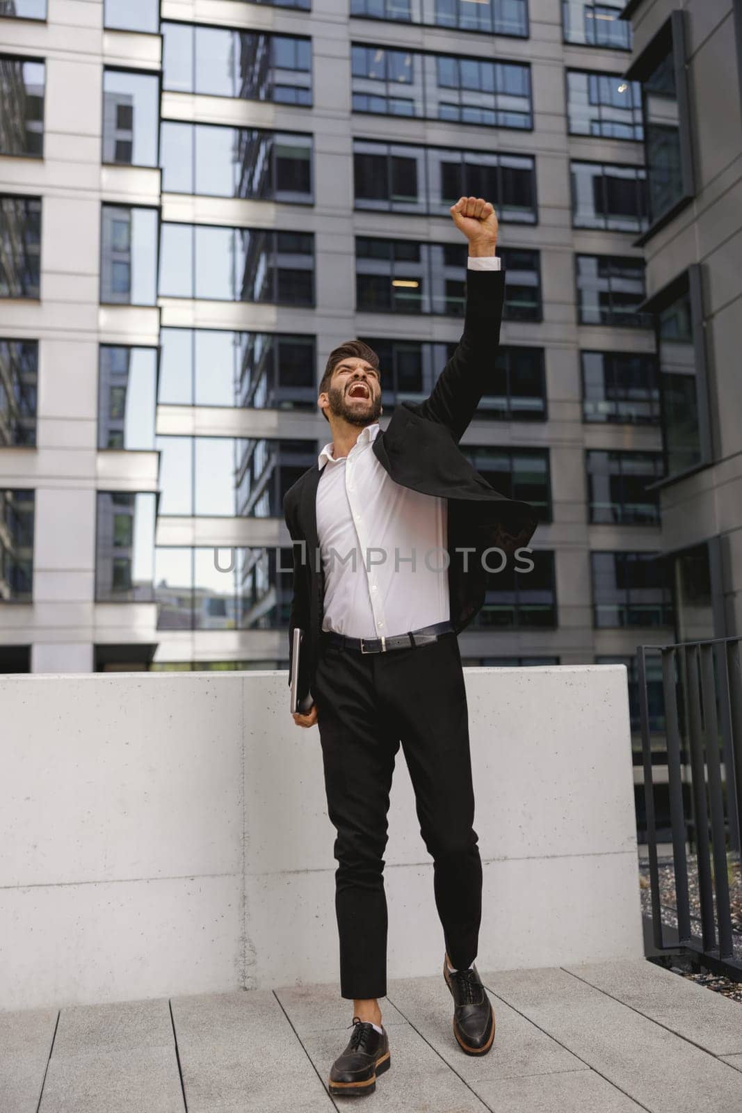 Businessman with laptop standing on office terrace and raised fist is suttisfied of finished project by Yaroslav_astakhov