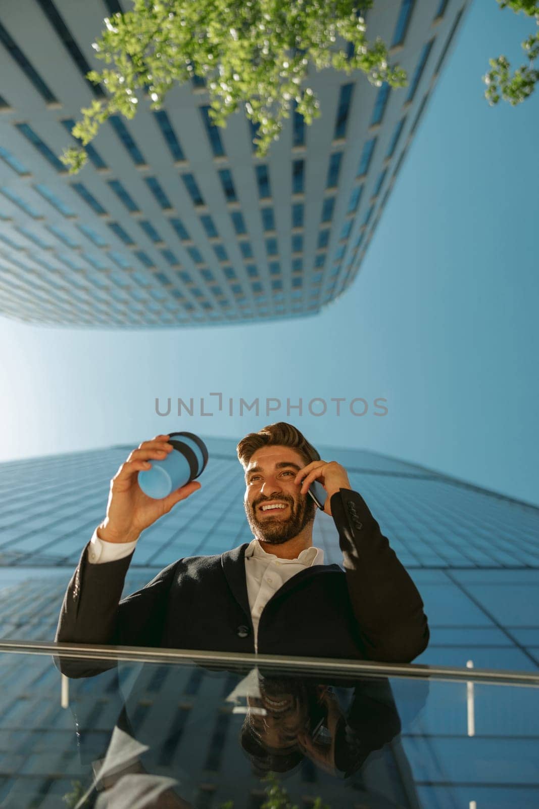 Male manager is talking phone and drinking coffee standing on background of city skyscrapers
