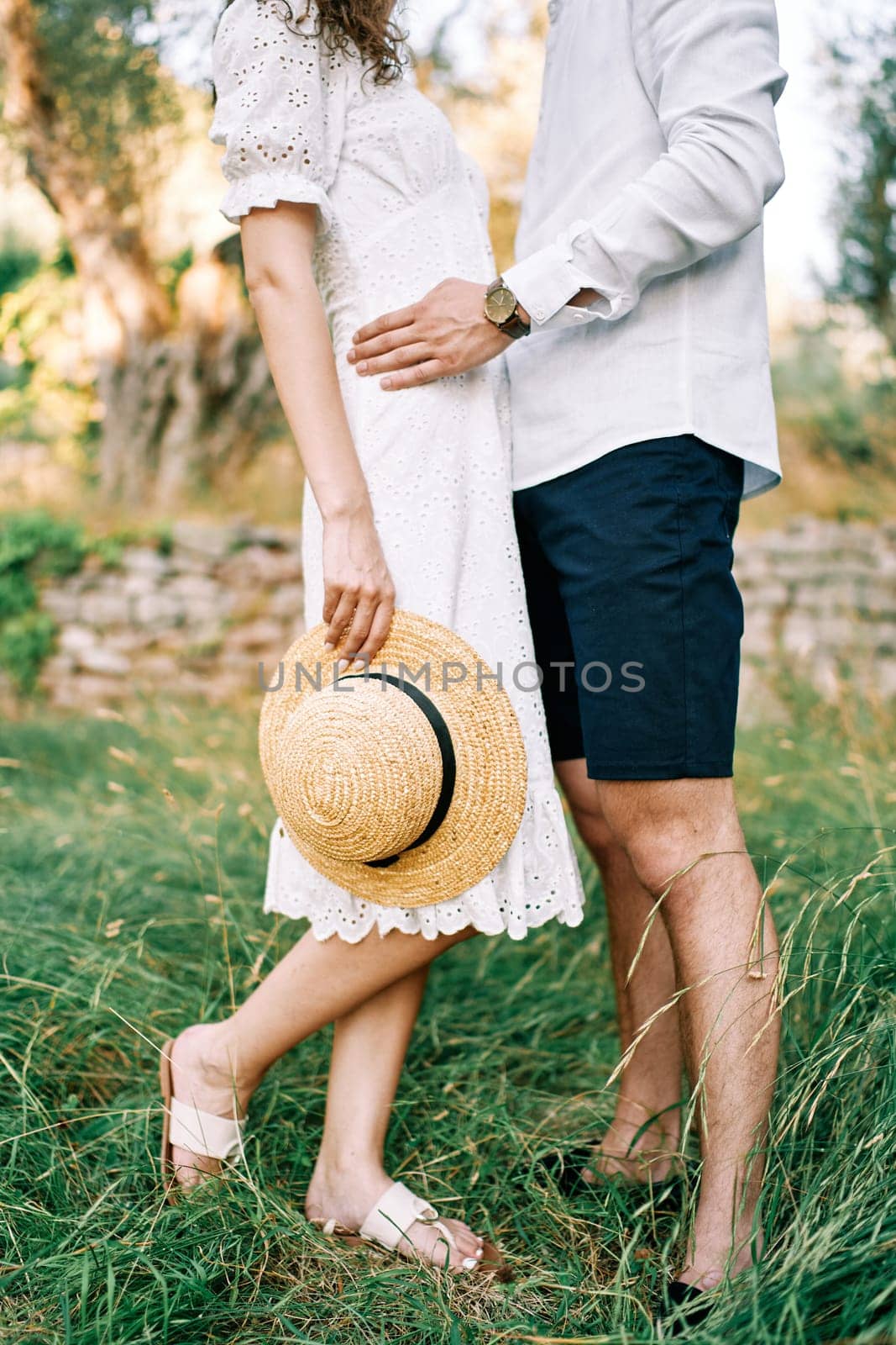 Man hugs woman by the waist standing with a straw hat in her hand in the garden. Cropped. Faceless by Nadtochiy