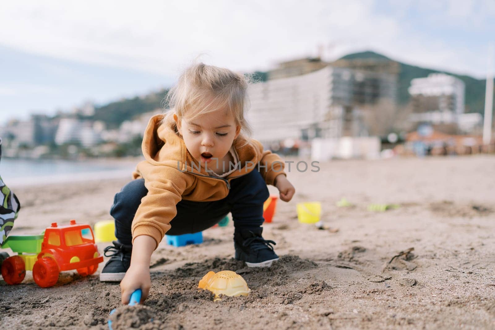 Little girl squats on the beach and digs the sand with her mouth open. High quality photo