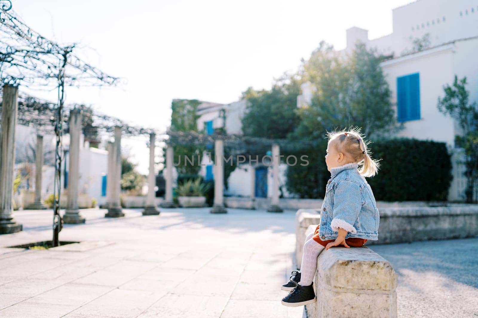 Little girl sits on a stone fence near the house and looks at the pergola in the garden. Side view. High quality photo