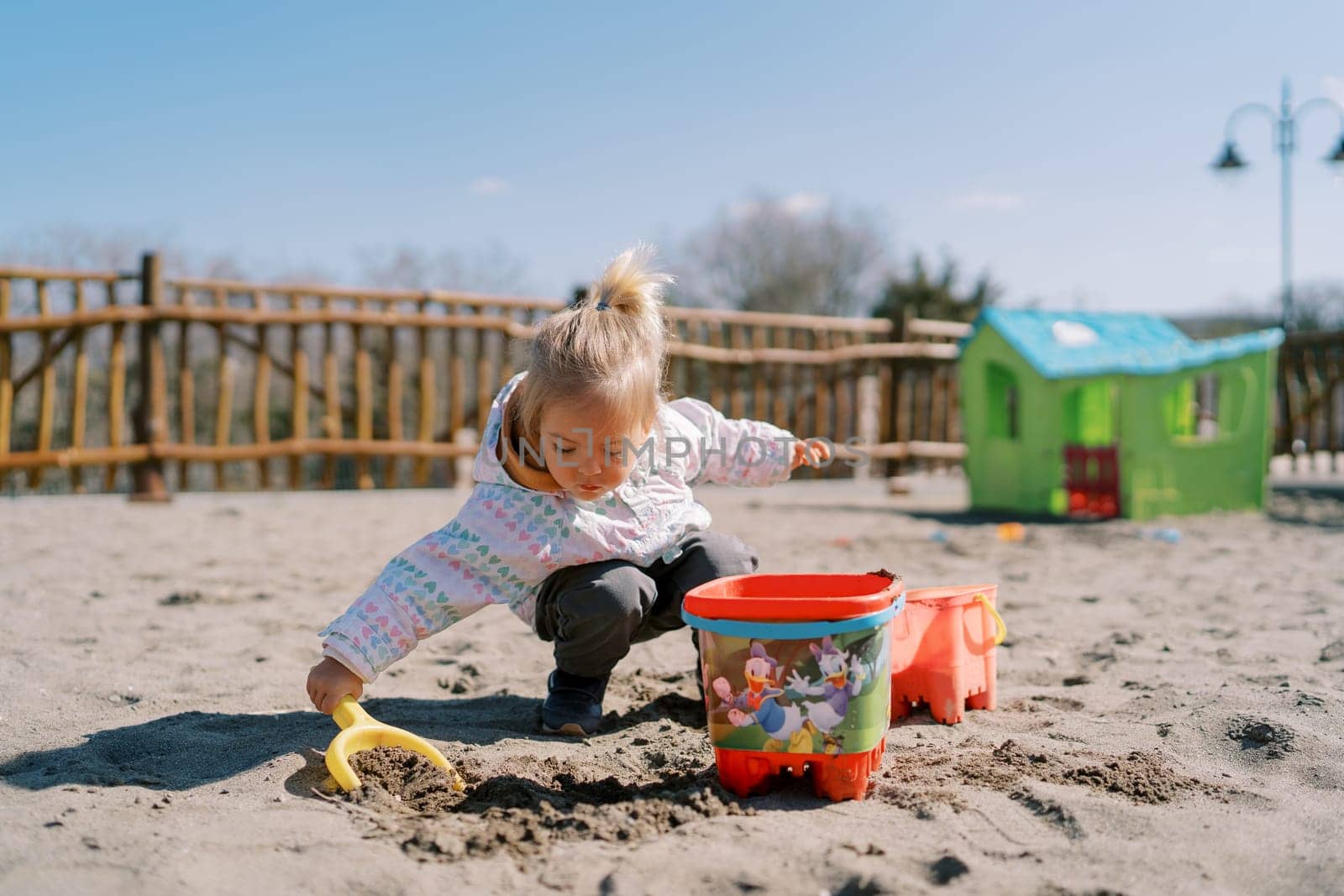 Little girl digs sand with a plastic shovel at the playground and pours it into a toy bucket by Nadtochiy