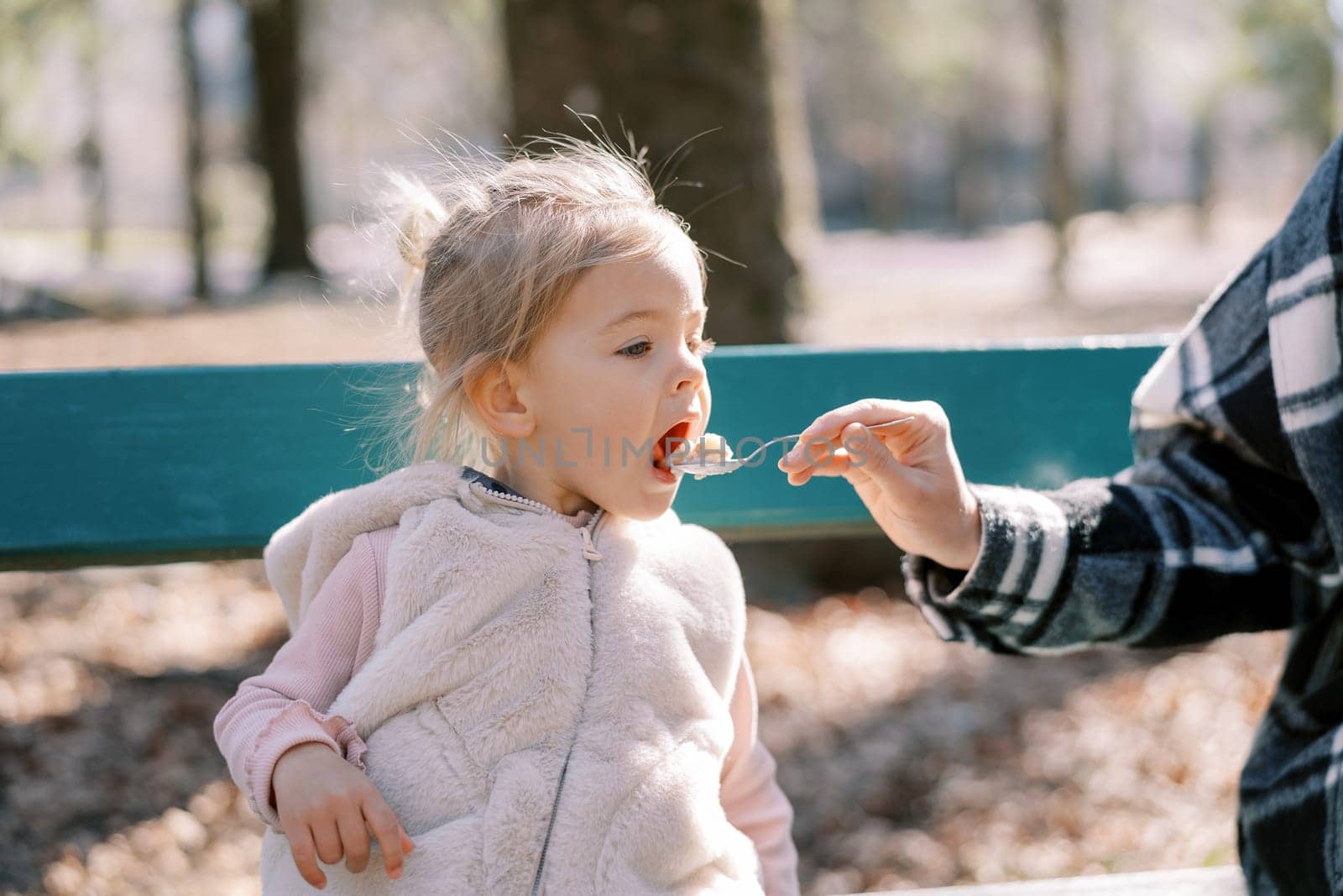 Mom feeds porridge from a spoon to a little girl sitting on a bench in the park. Cropped. High quality photo