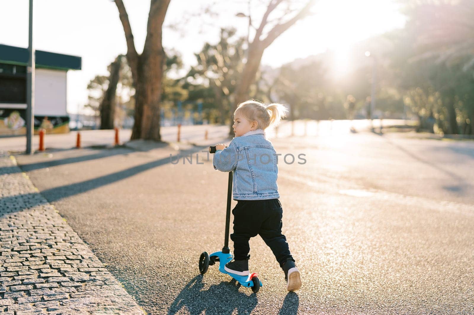 Little girl rides a scooter on an asphalt road and looks away. Back view. High quality photo