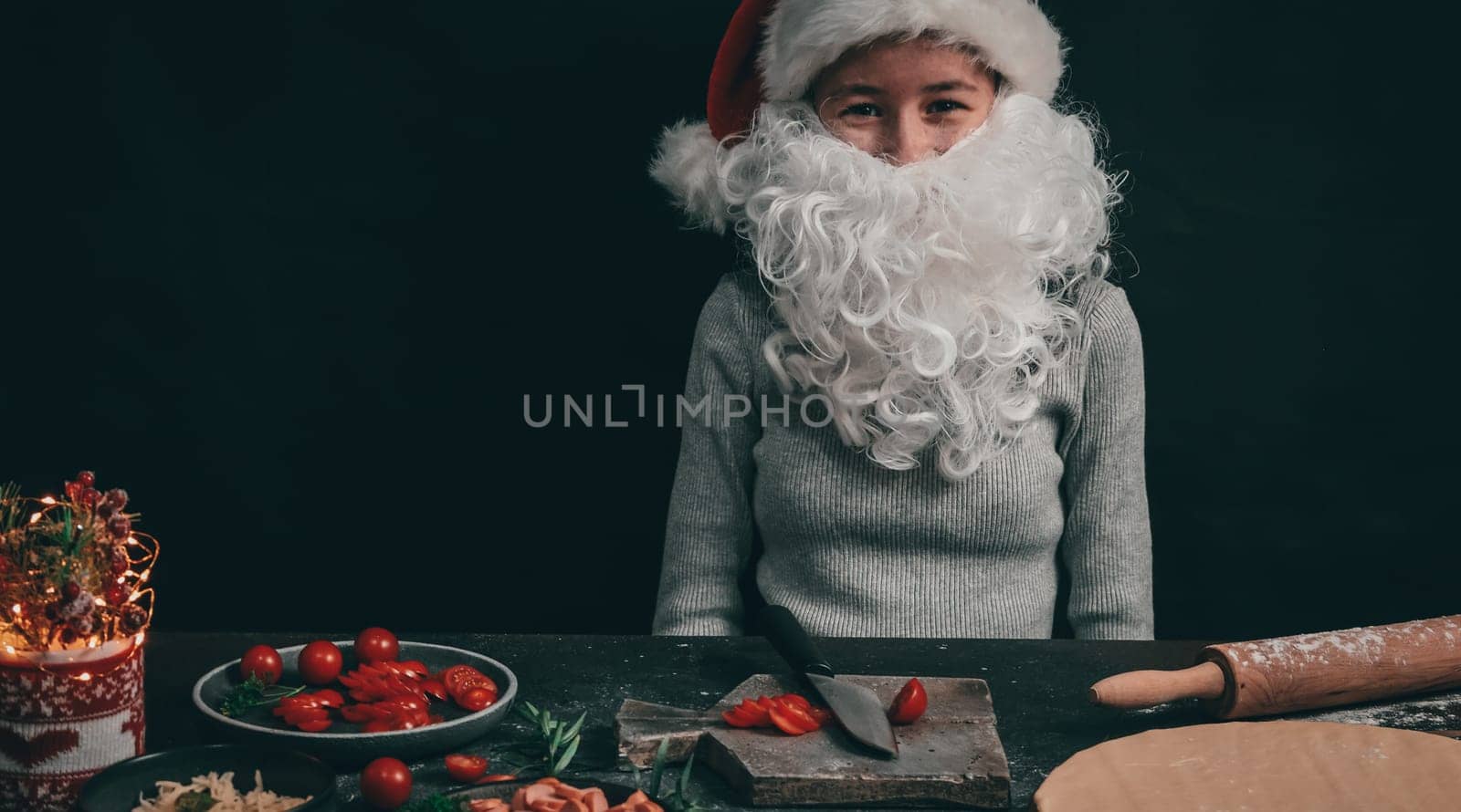 Caucasian teenage girl in a Christmas hat with a beard sits cheerful at the table with cherry tomatoes on a gray wooden board, next to pizza dough, rolling pin and Christmas decor on a dark background, close-up side view. Cooking concept.