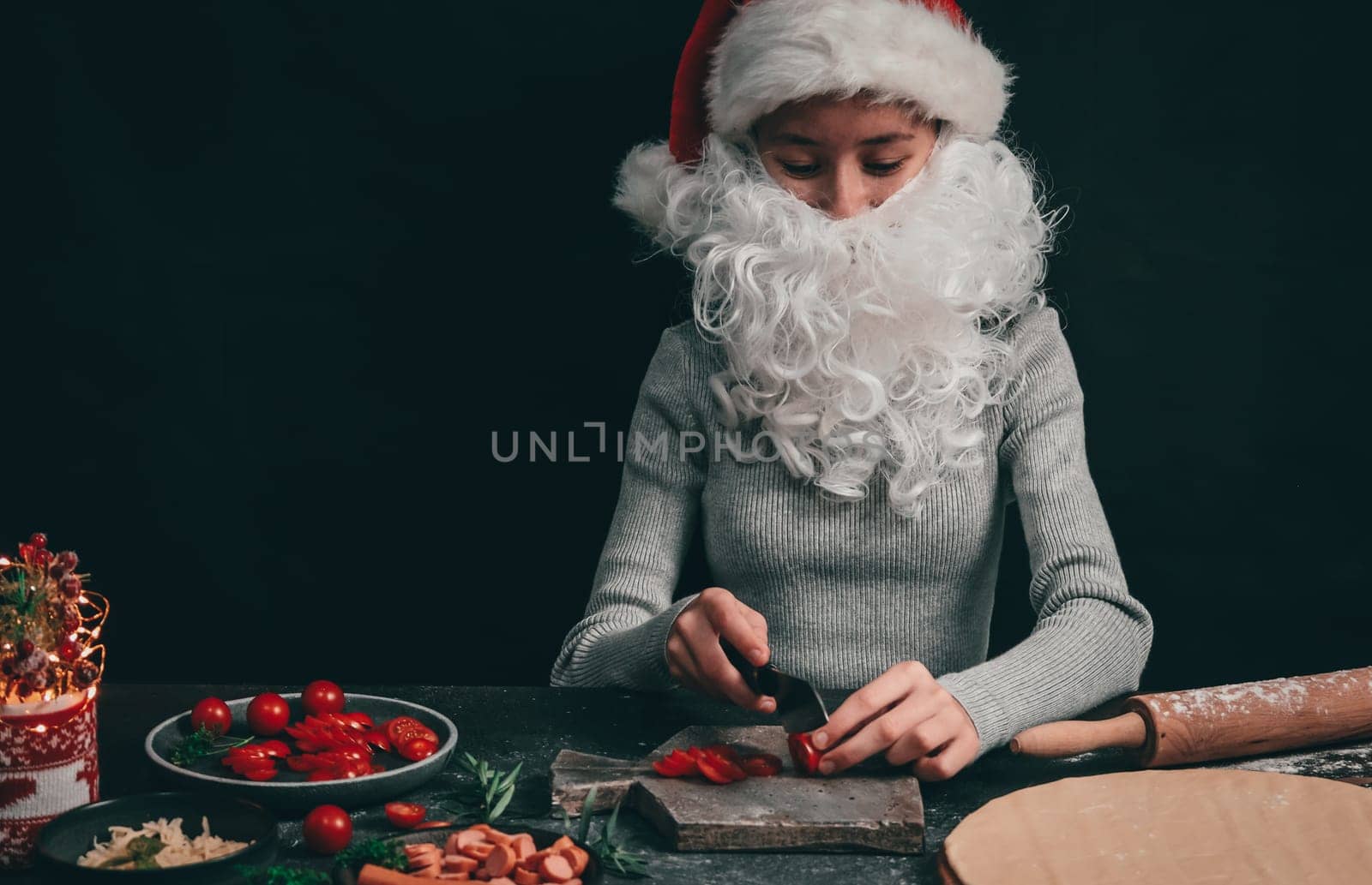 Caucasian teenage girl in a santa hat with a beard cuts with a big black knife cherry tomatoes on a gray wooden board, next to pizza dough, rolling pin and Christmas decor on a dark background, close-up side view. Cooking concept.