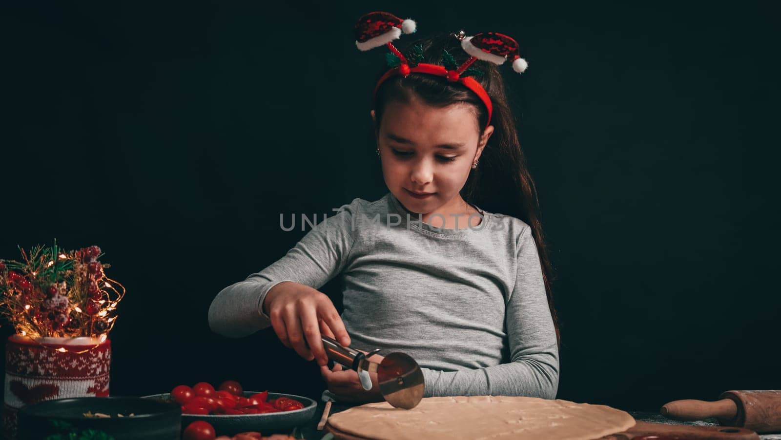 A Caucasian little girl cuts a Christmas tree with a round moon knife on a pizza dough, and next to a rolling pin, sliced cherry tomatoes, ketchup and a decoration in a mug with a sweater on a dark background, close-up side view.