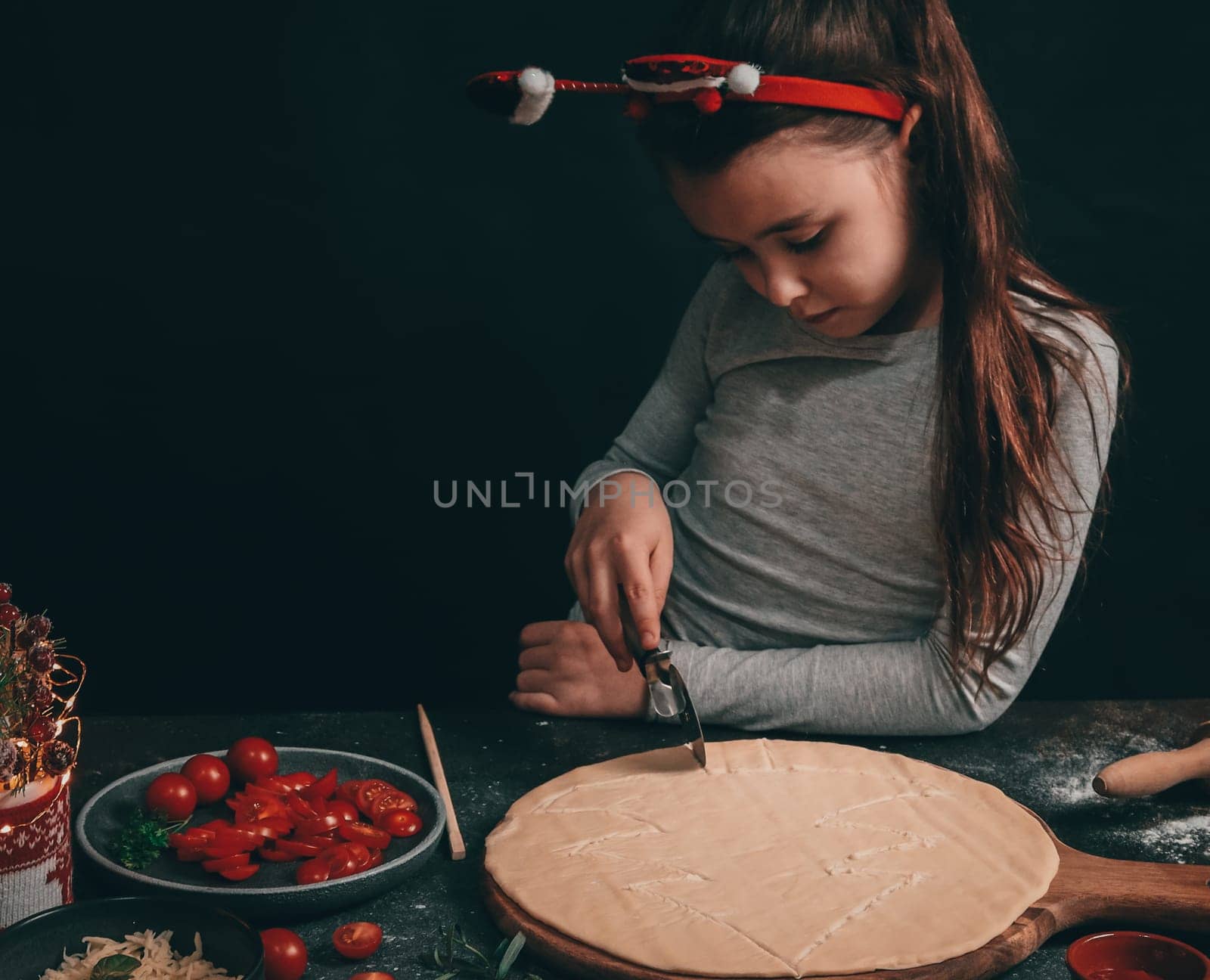 A Caucasian little girl cuts a Christmas tree with a round moon knife on a pizza dough, and next to a rolling pin, sliced cherry tomatoes, ketchup and a decoration in a mug with a sweater on a dark background, close-up side view.