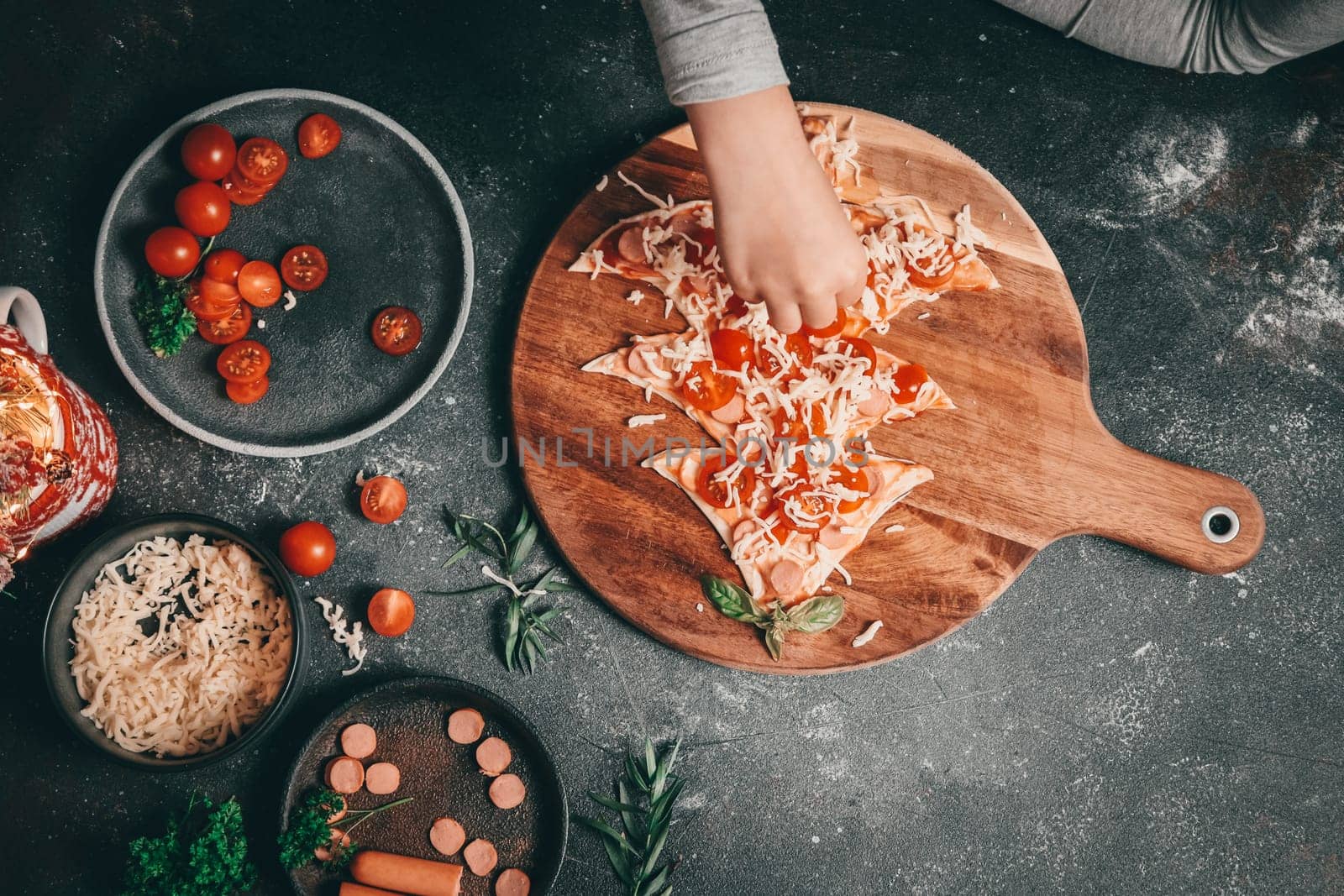 Children hand sprinkles mozzarella cheese on a Christmas tree cut from pizza dough on a cutting board lies on a dark background with cherry tomatoes, sausages and decoration, top view close-up. The concept of preparing for Christmas and New Year.