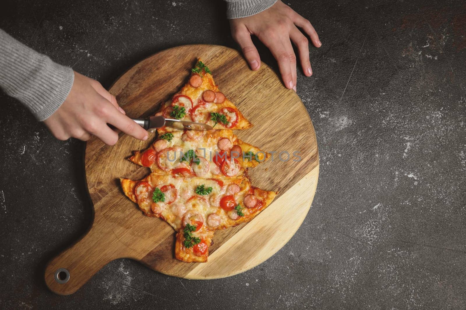 Hands of a Caucasian teenage girl cuts a moonknife pizza in the shape of a Christmas tree on a cutting board on a dark background, close-up top view.